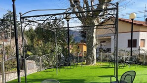 A patio with round tables and chairs, a tree, and a building in the background. - Hotel Caroline (Brusimpiano)