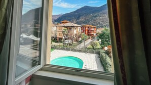A window with a view of a pool, buildings, and mountains. - Hotel Caroline (Brusimpiano)