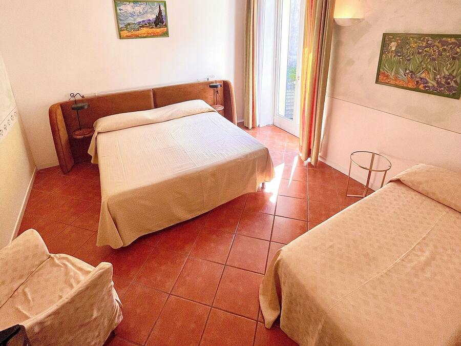 A hotel room with a large bed, a smaller bed, and a sofa. There are two paintings on the walls and a window with curtains.