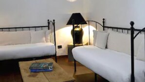 Una habitación pequeña y sencilla con dos camas individuales, una mesita de noche con lámpara y una mesa de madera.