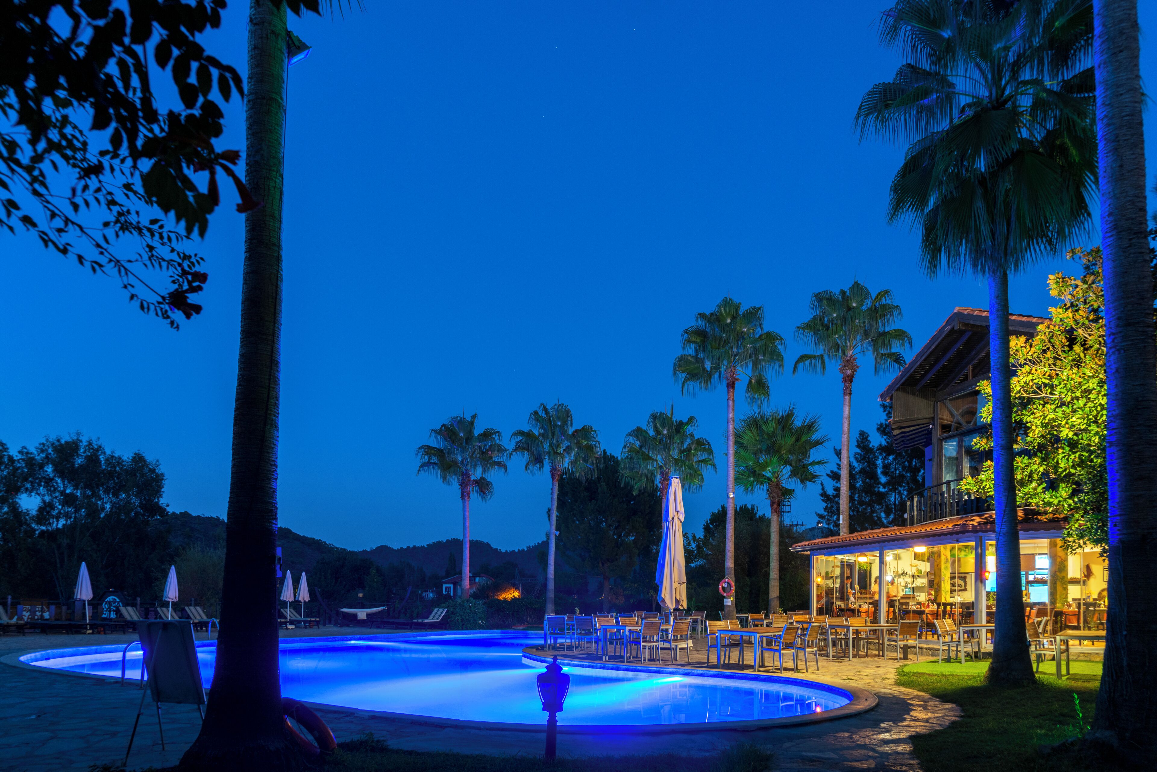 A pool area with lounge chairs and umbrellas, surrounded by palm trees and a building with outdoor seating.