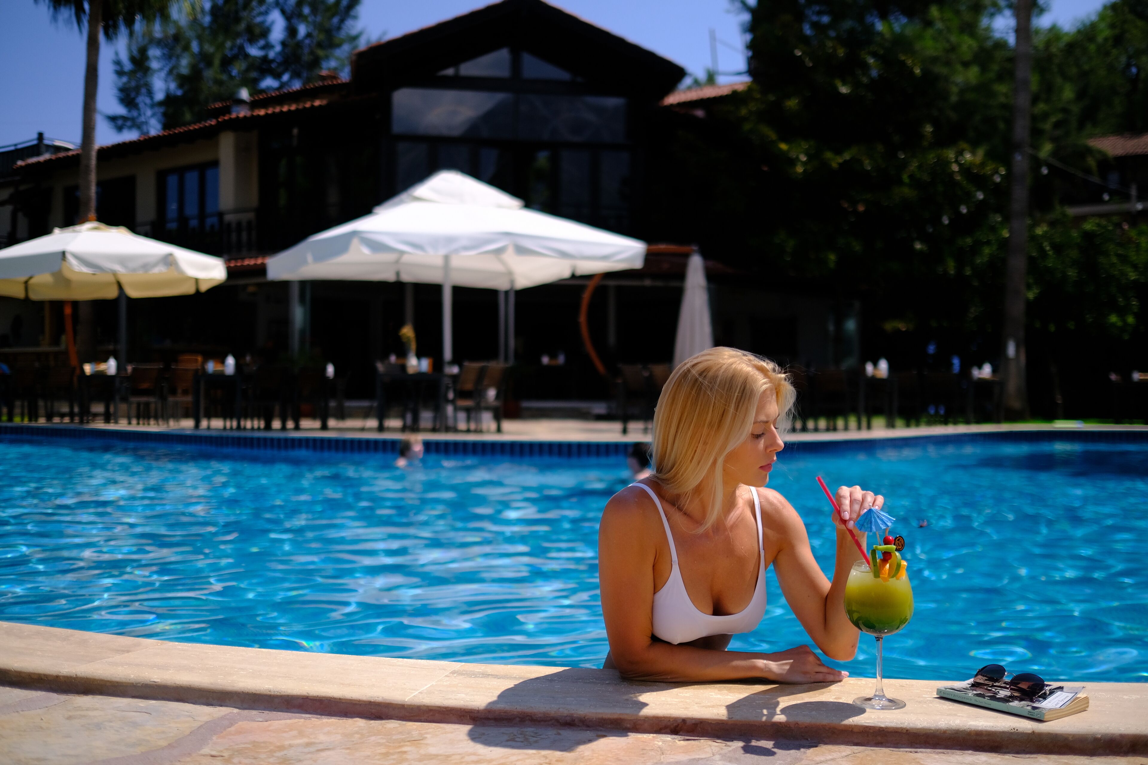 Escena junto a la piscina con una persona sosteniendo un cóctel, un vaso y gafas de sol sobre una mesa.