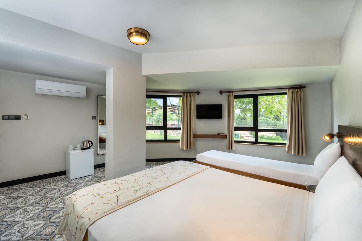 A hotel room with a bed, a small desk with a kettle, an air conditioner, a TV, and windows with curtains.