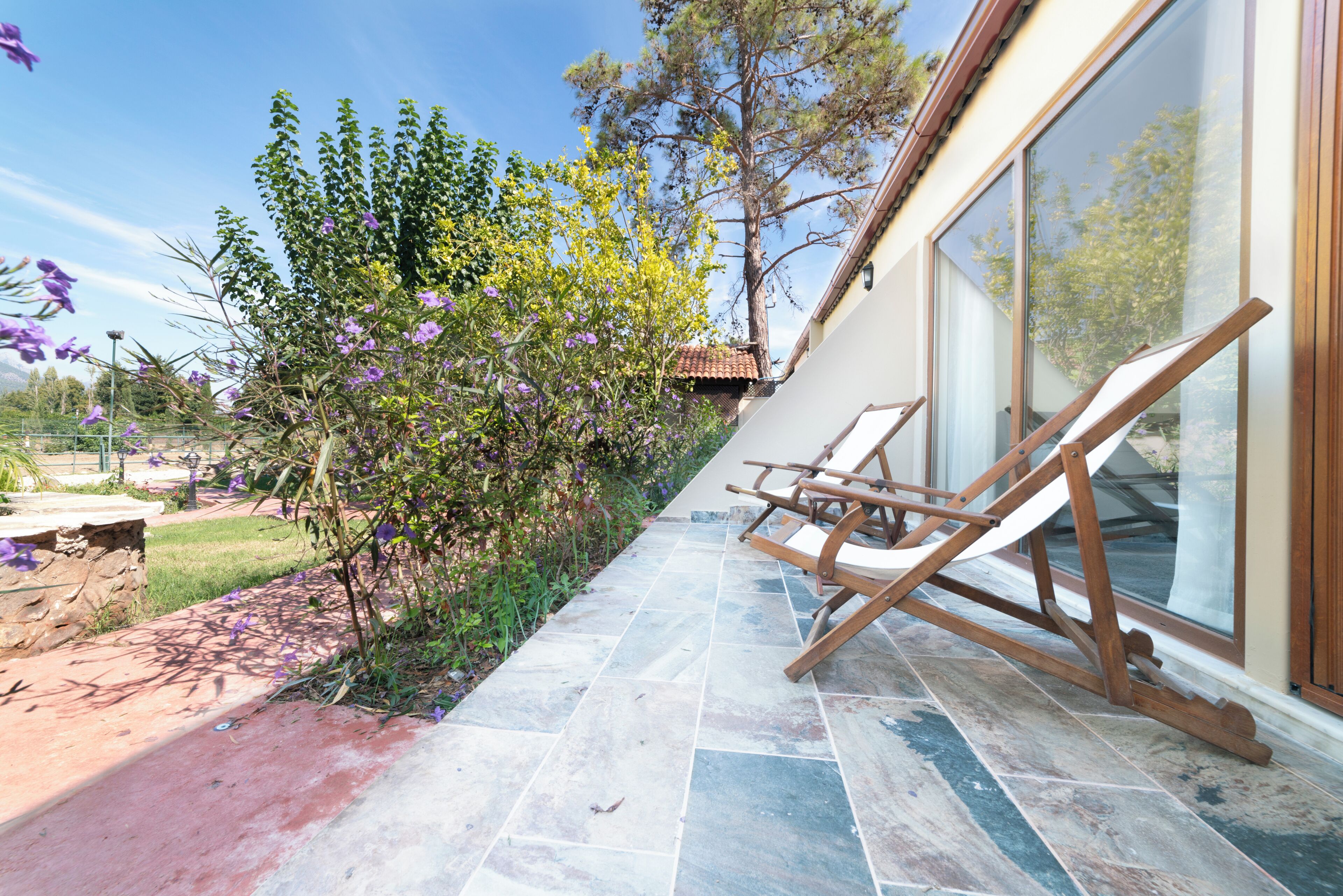 A patio with a stone floor, a wooden swing chair, and a glass door leading to a garden with trees and purple flowers.