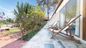 A patio with a stone floor, a wooden swing chair, and a glass door leading to a garden with trees and purple flowers.