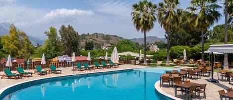 Zona de piscina con sillones de playa y sombrillas, rodeada de palmeras y montañas al fondo.