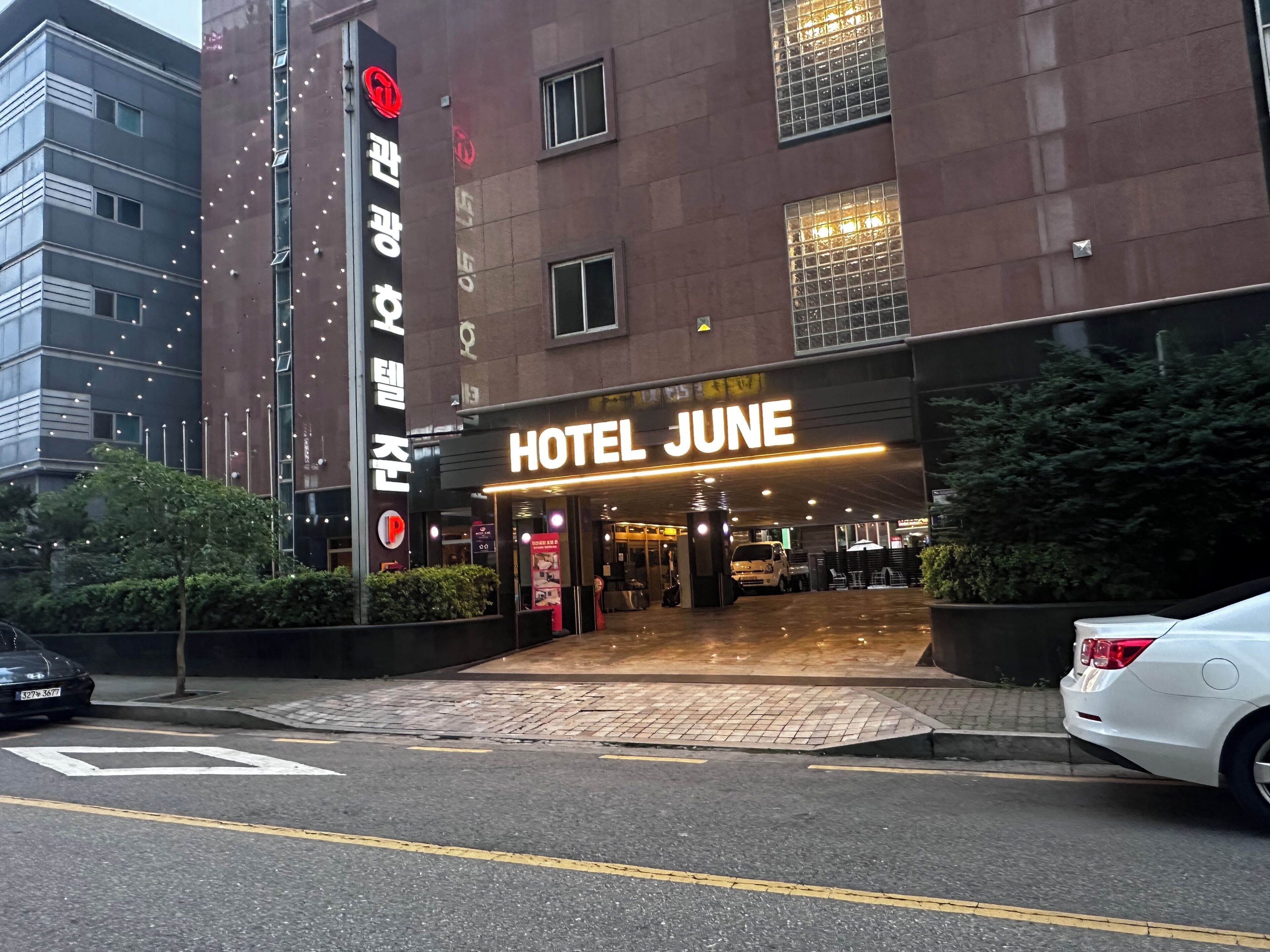 A hotel entrance with a sign reading 'HOTEL JUNE,' a parking lot, and a building with a red logo.