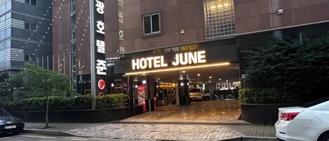 A hotel entrance with a sign reading 'HOTEL JUNE,' a parking lot, and a building with a red logo.