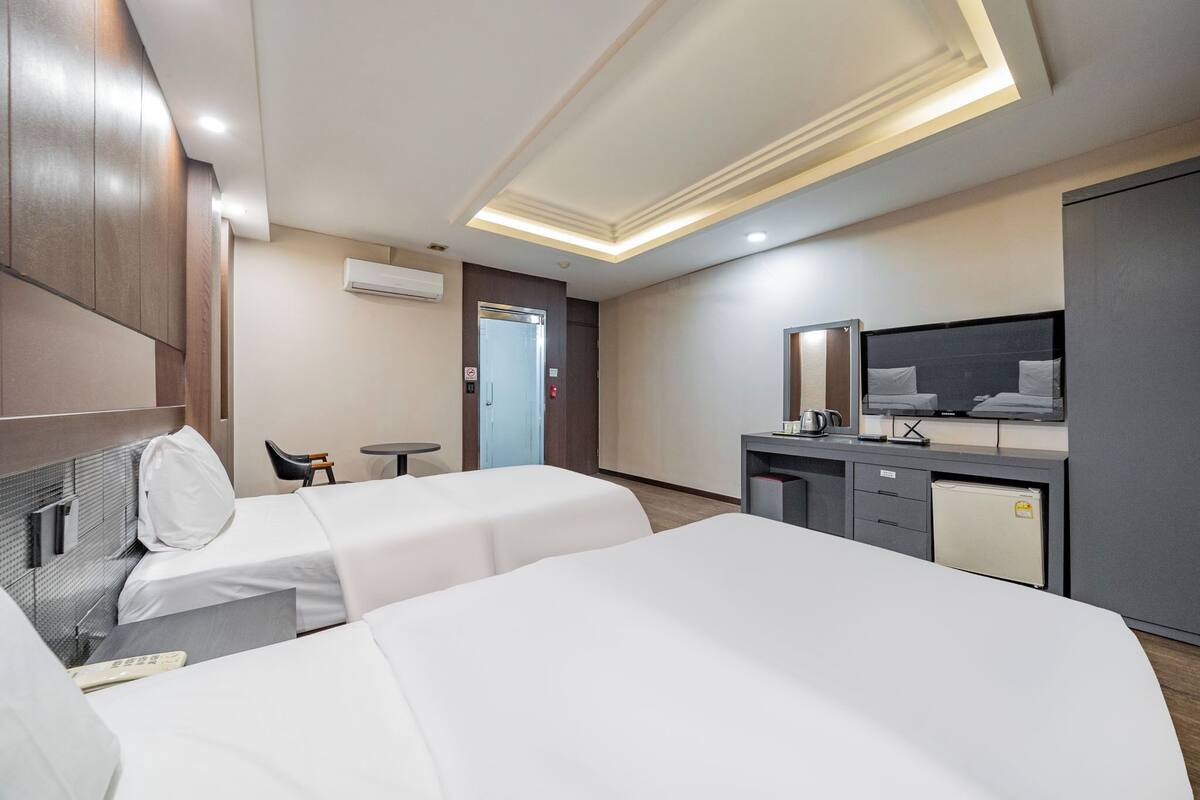 A hotel room with two beds, a small table, a chair, a TV, and a mini-fridge.