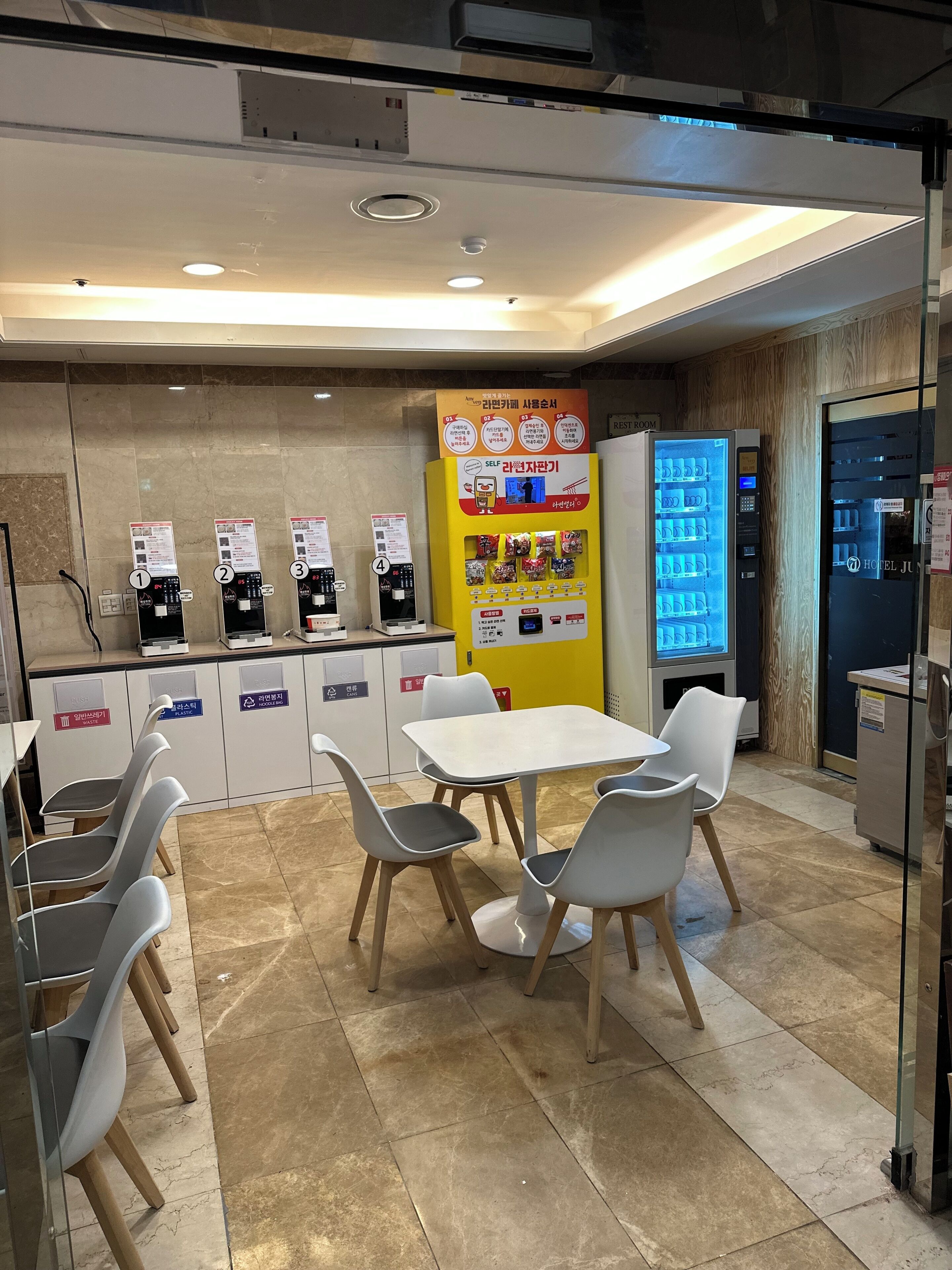 A hotel lobby with a self-service beverage machine, a rest room sign, a vending machine, and a seating area with chairs and a table.