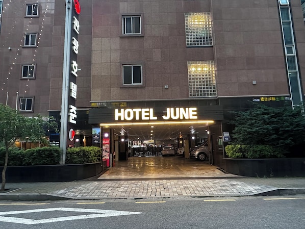A hotel entrance with a sign reading 'HOTEL JUNE,' a parking area, and a building facade with windows.