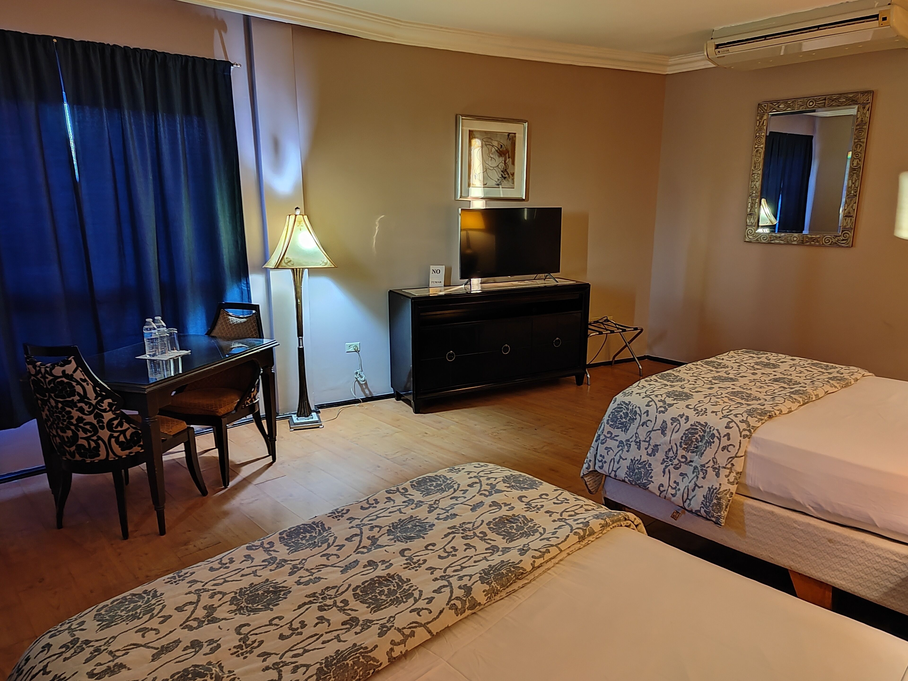 A hotel room with two beds, a desk, a chair, a TV, a lamp, and a mirror.