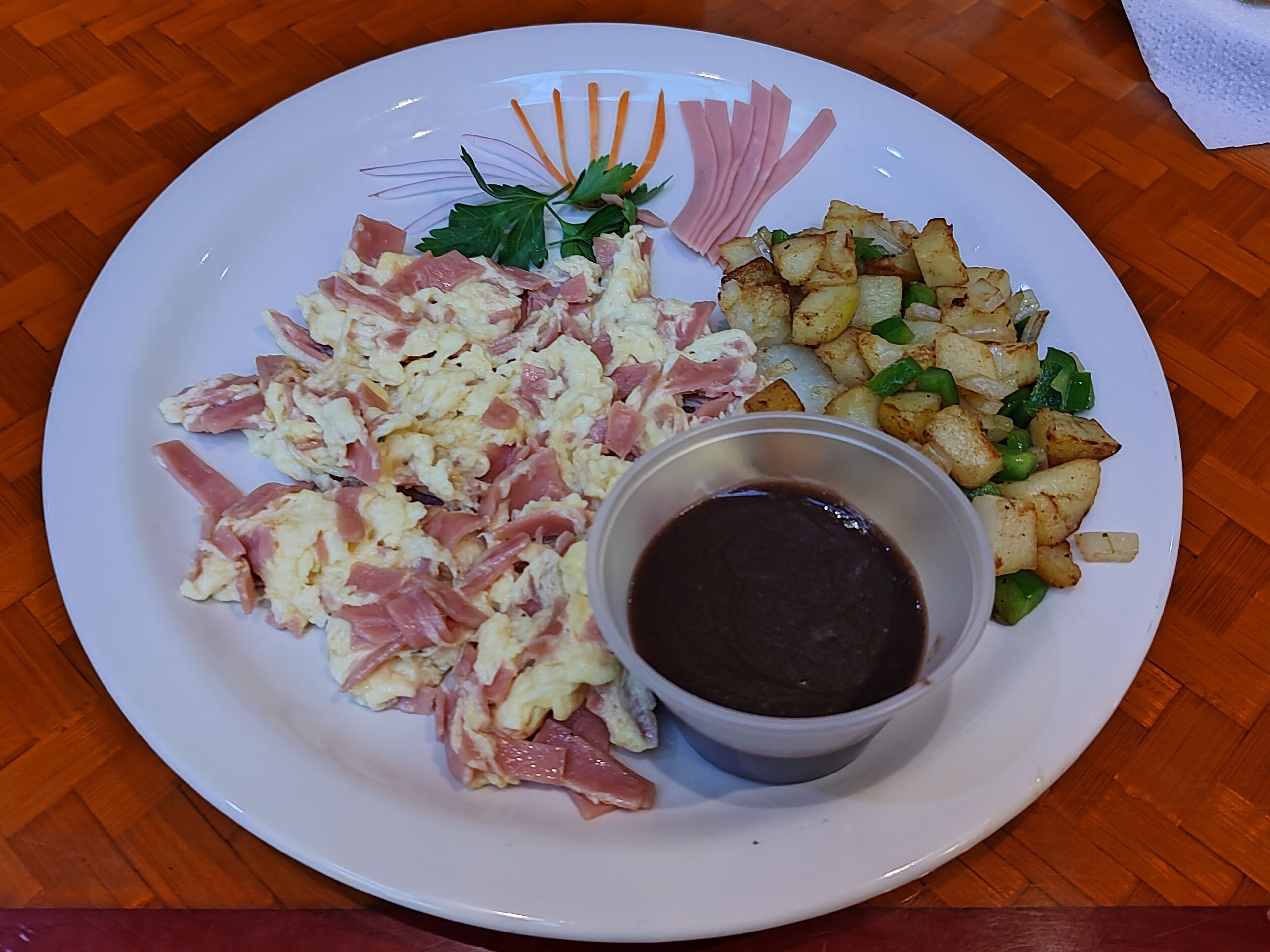 Un plato con huevos revueltos, jamón, patatas asadas y un recipiente pequeño con salsa.