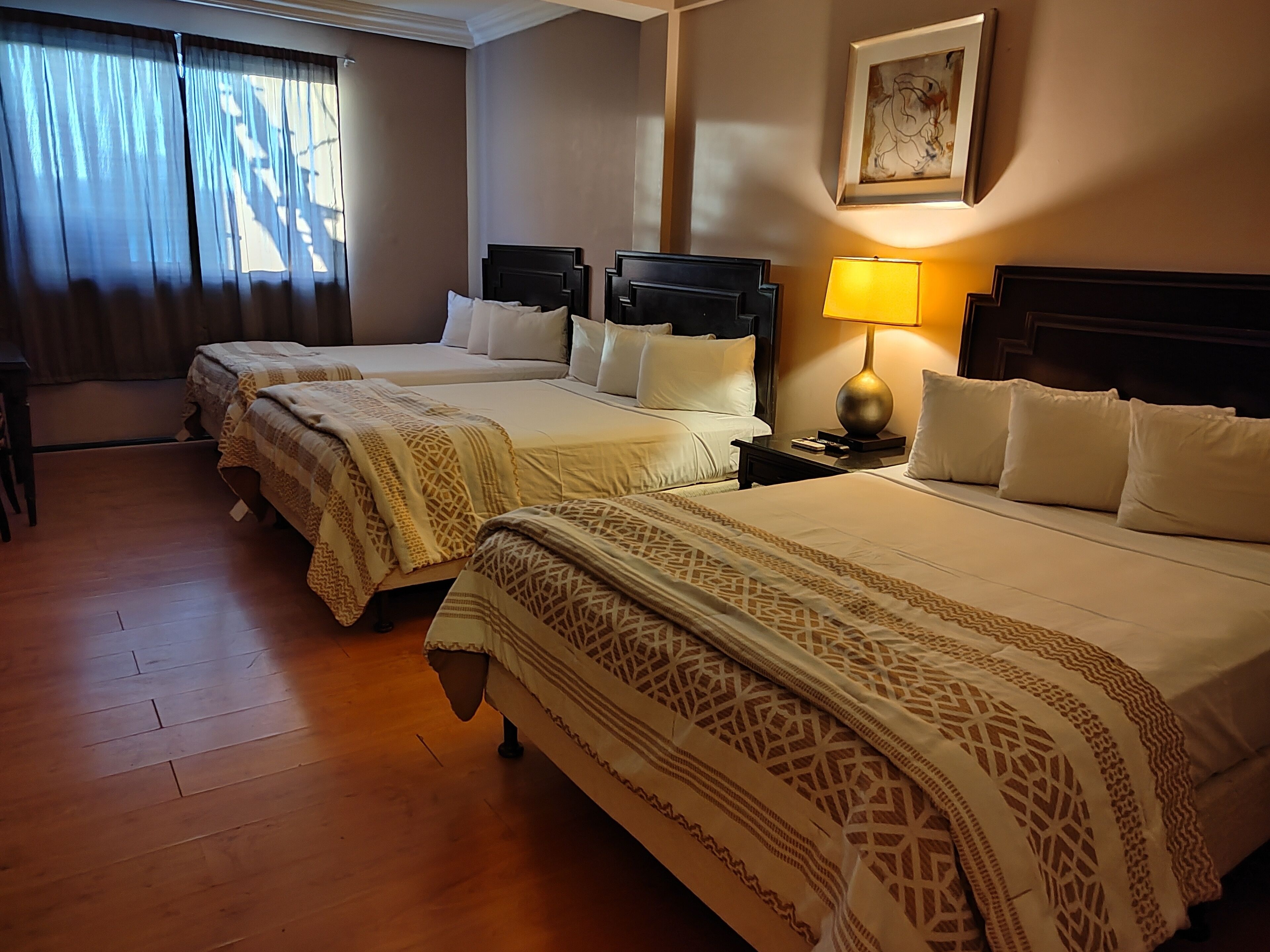 Habitación de hotel con tres camas, piso de madera, una mesita de noche con lámpara y un cuadro enmarcado en la pared.