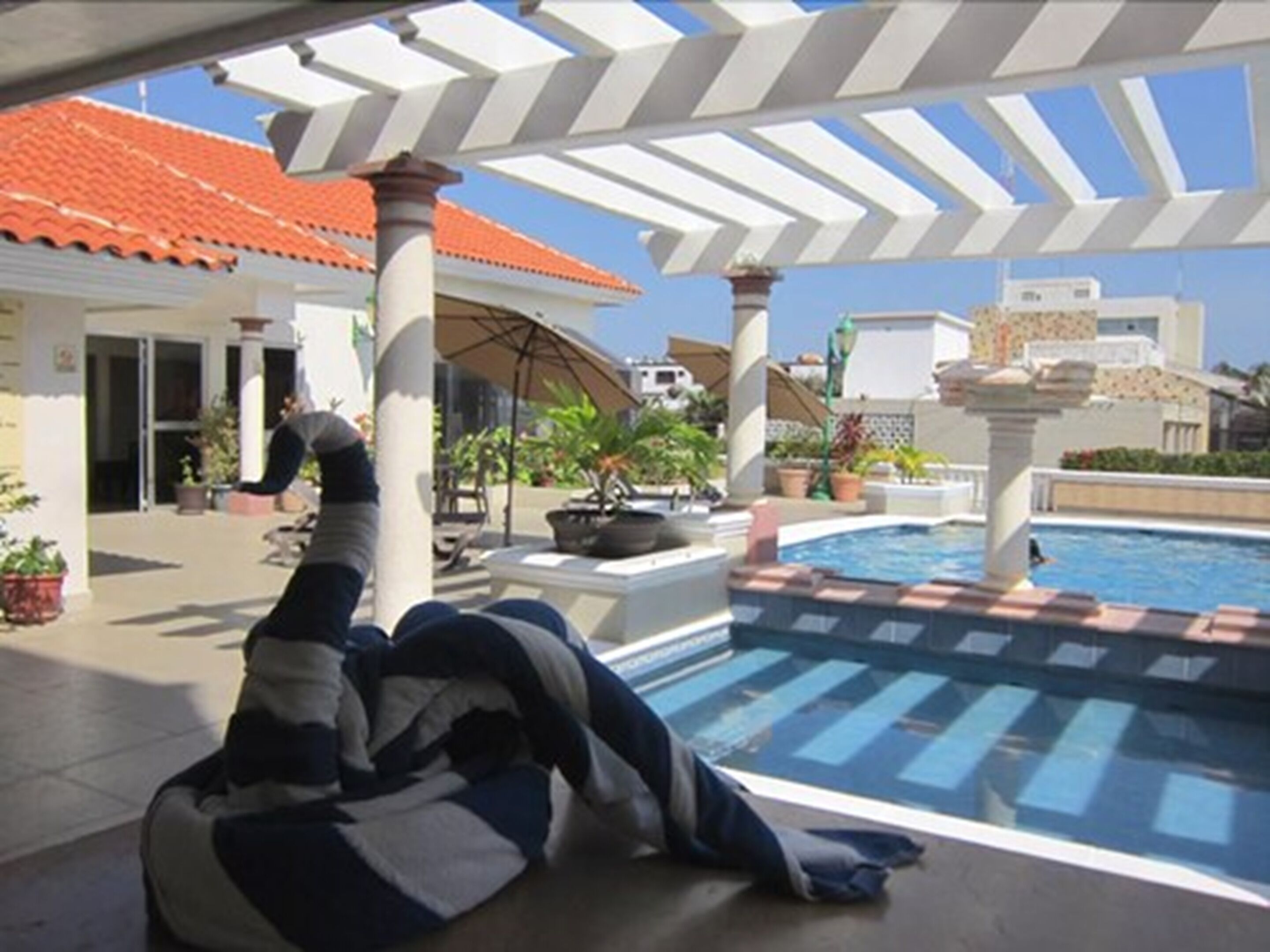 Une zone avec une piscine où l’on voit une personne allongée au bord, un patio couvert avec des colonnes, et un bâtiment avec un toit en tuiles en arrière-plan.
