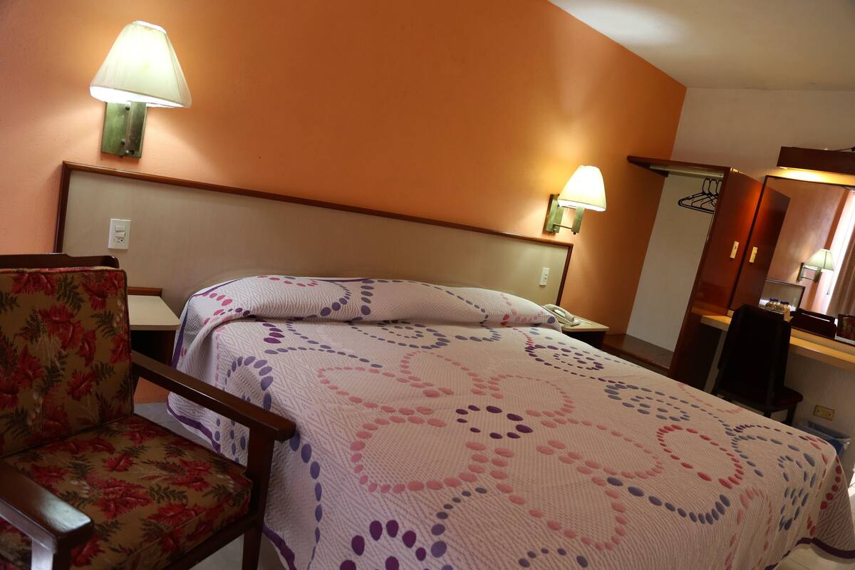 Quarto de hotel com cama, cadeira, mesa de cabeceira e abajur.