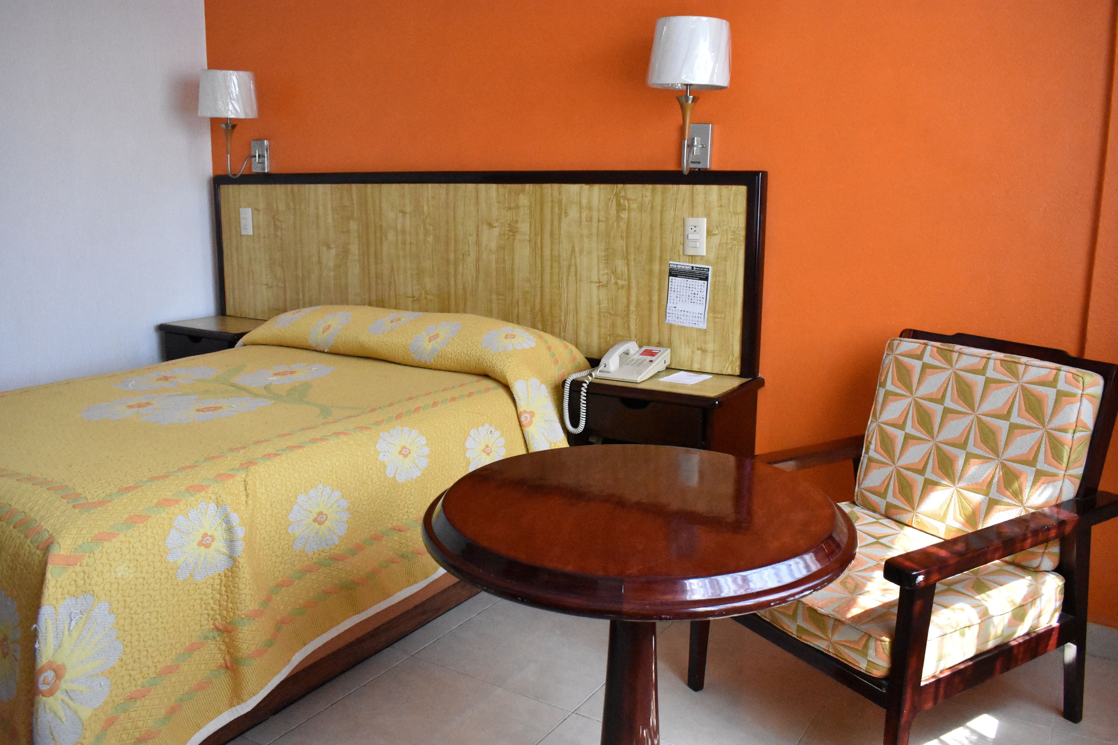A hotel room with a bed, a bedside table with a lamp, a chair, and a small round table.