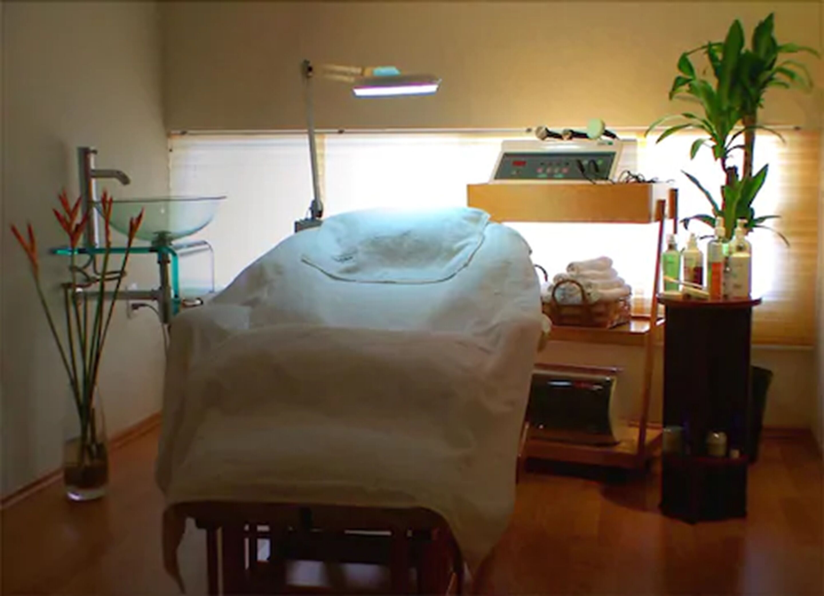 Una camera con un letto coperto da un lenzuolo bianco, una scrivania con un computer, una lampada, una pianta e uno scaffale con articoli da toilette.