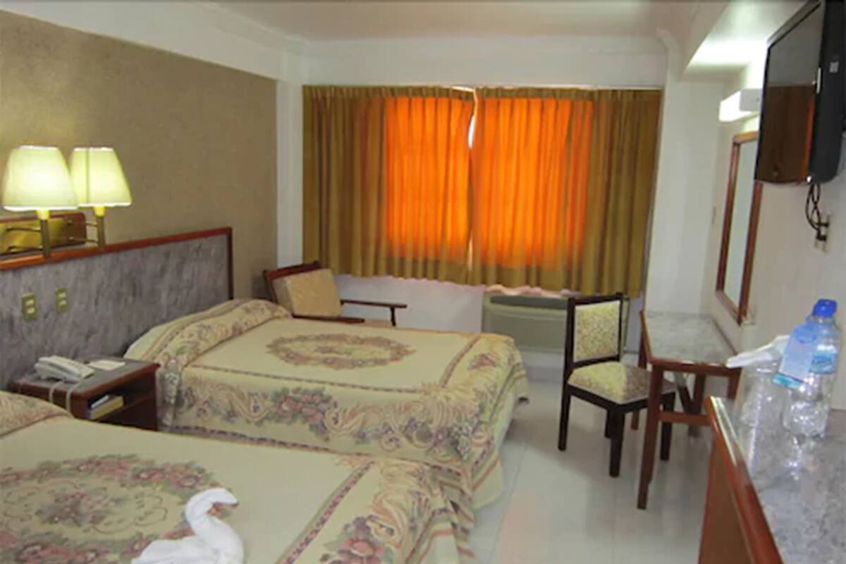 Quarto de hotel com duas camas, uma escrivaninha, uma cadeira, uma televisão e um banheiro.
