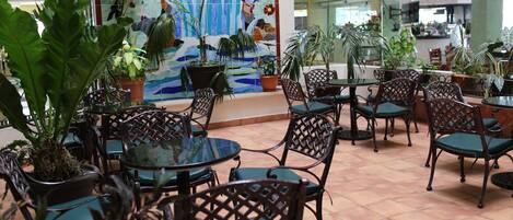 Un espacio interior con mesas y sillas, rodeado de plantas en macetas y un mural colorido.