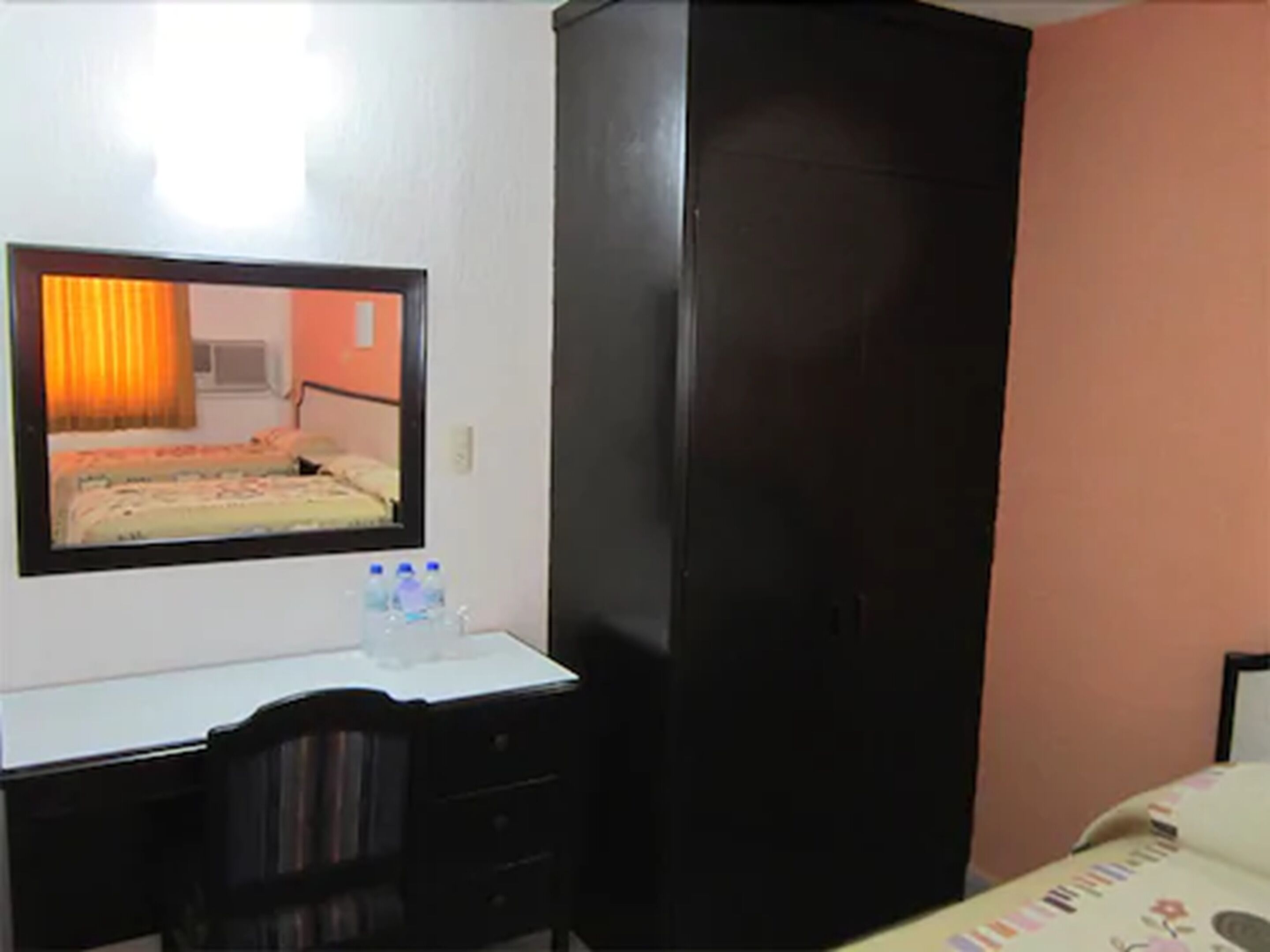 A hotel room with a bed, a desk with a mirror, a wardrobe, and a bedside table with water bottles.