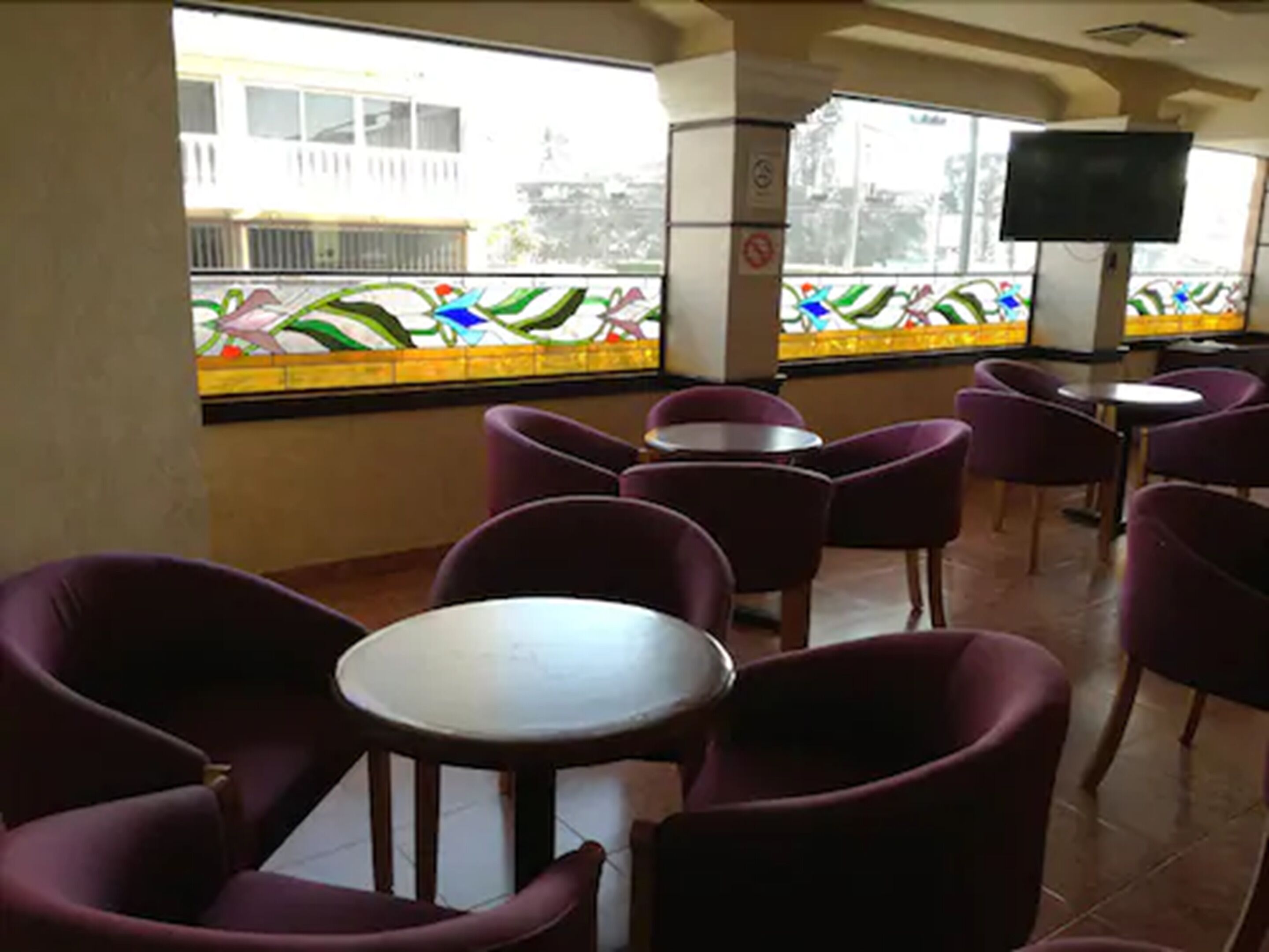 A restaurant interior with purple chairs and tables, a large window with a colorful stained glass design, and a television mounted on the wall.