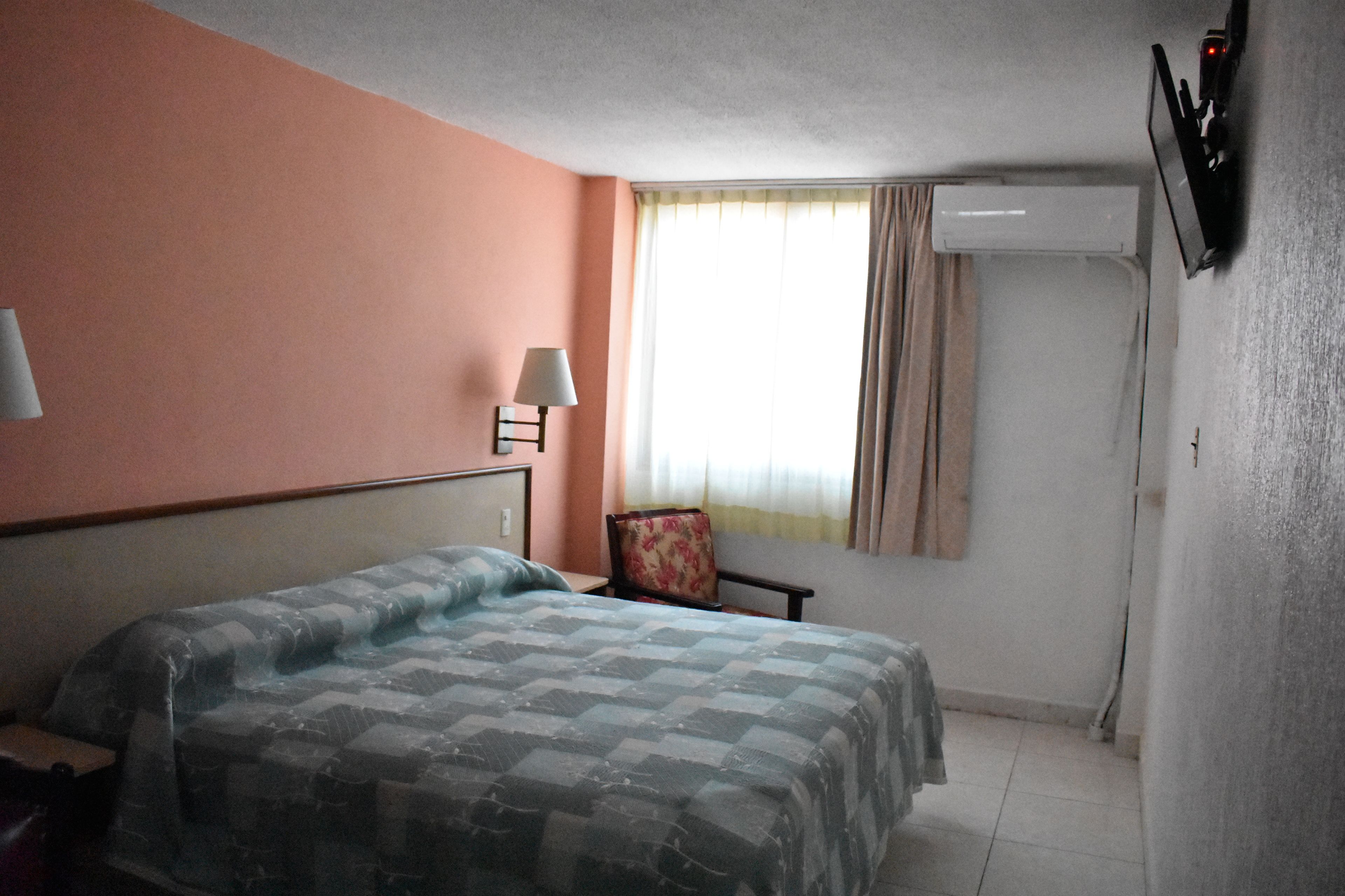 A hotel room with a bed, a chair, a lamp, and a window with curtains.