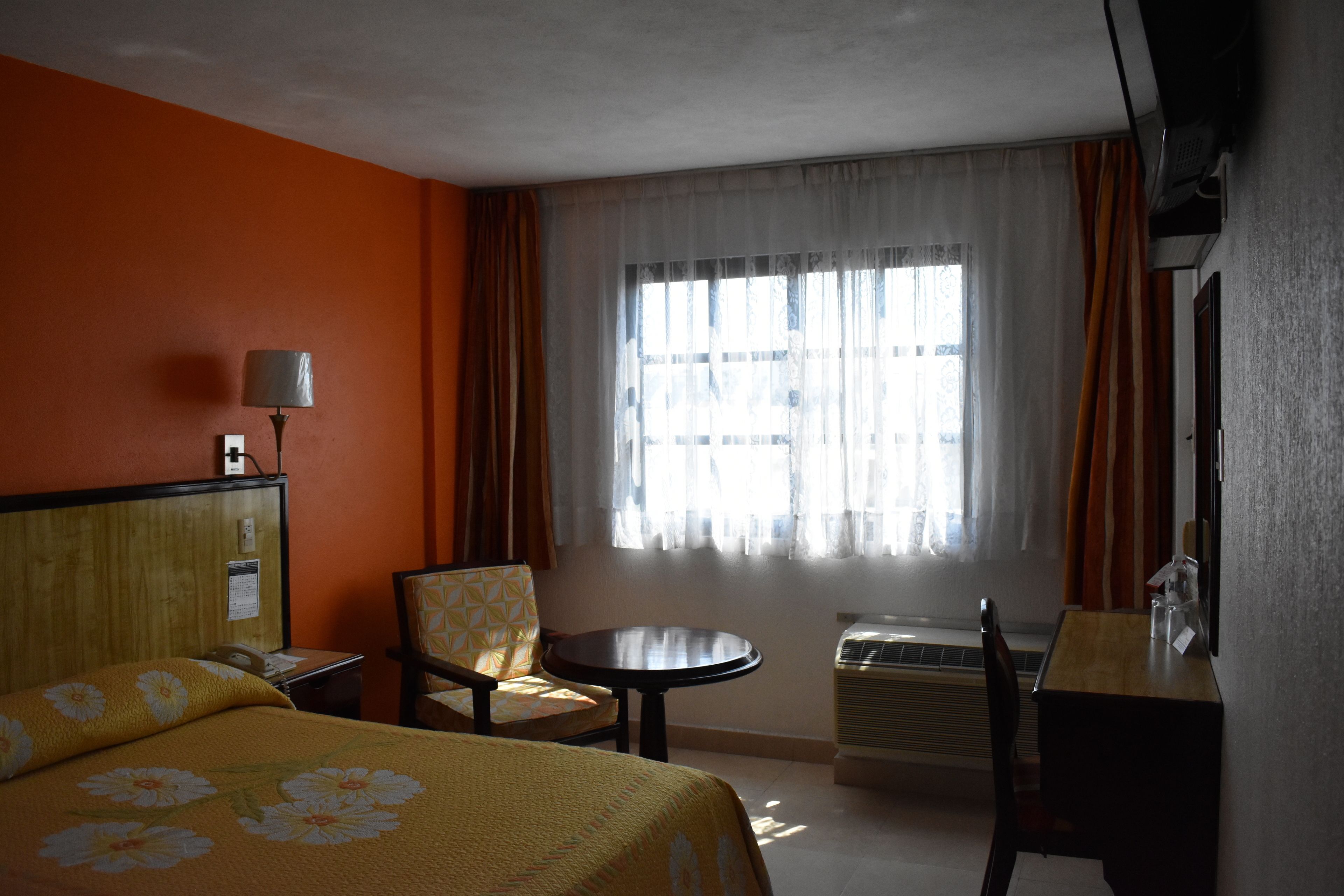 Una habitación de hotel con cama, una silla, una mesita, escritorio, lámpara y ventana con cortinas.