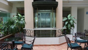 A hotel lobby with a central elevator, surrounded by outdoor seating with chairs and tables, and potted plants.