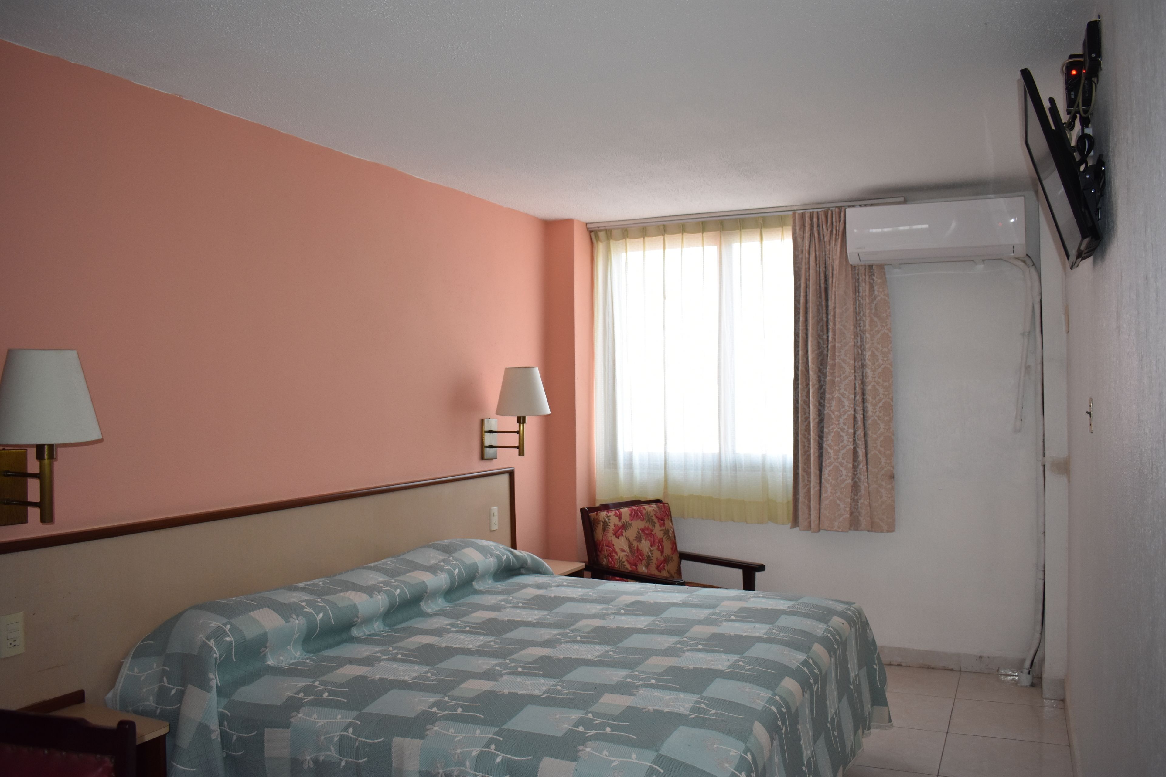 Habitación de hotel con cama, dos lámparas de noche, una silla, ventana con cortinas y aire acondicionado instalado.
