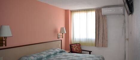 Habitación de hotel con cama, dos lámparas de noche, una silla, ventana con cortinas y aire acondicionado instalado.