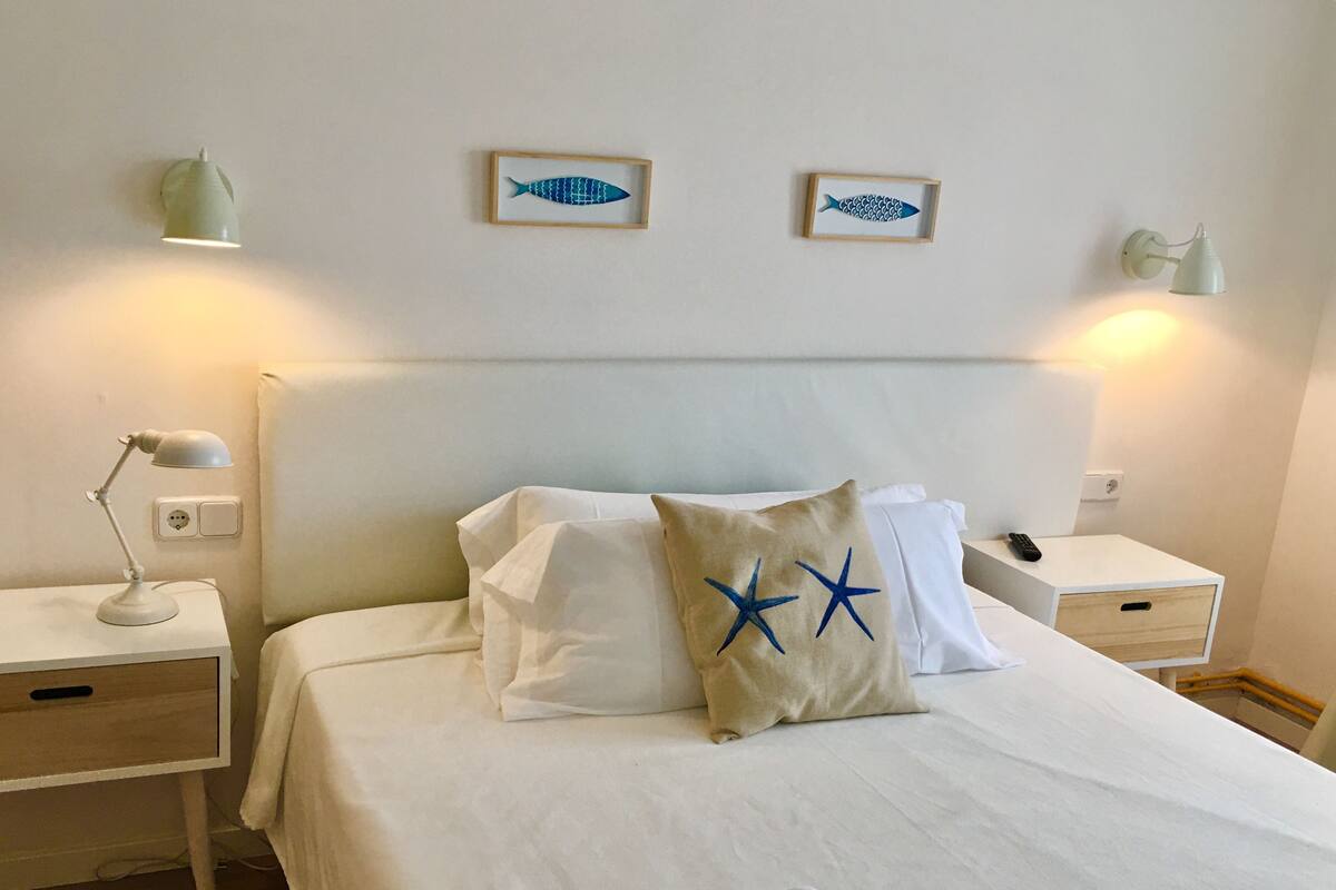 Una cama bien hecha con sábanas blancas, dos almohadas con diseños de estrellas, una lámpara de noche y una obra de arte con peces enmarcada en la pared.