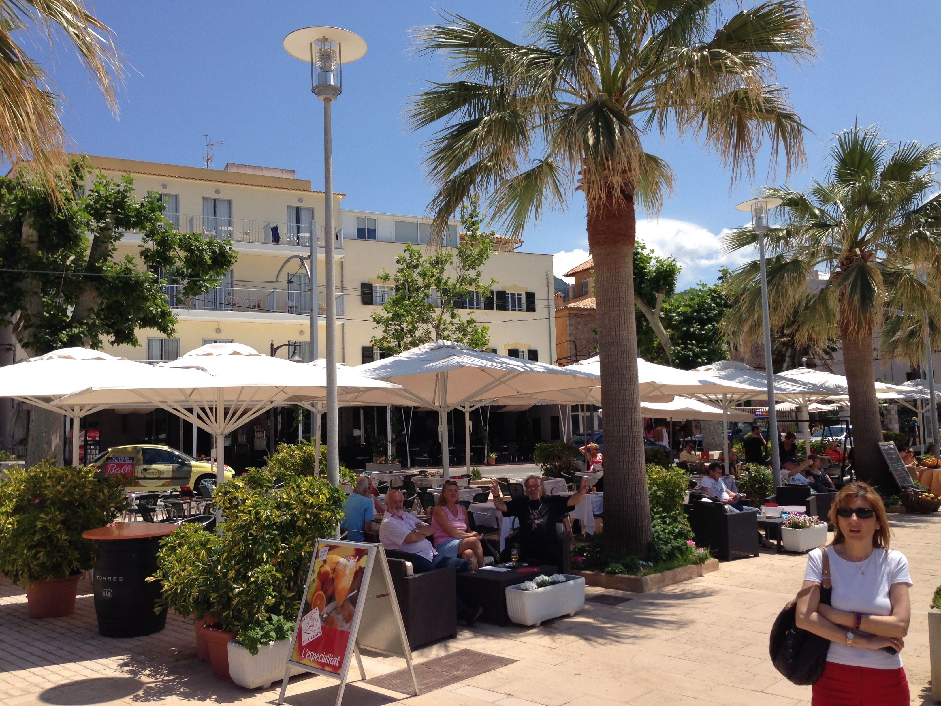 An outdoor café area with tables and chairs under umbrellas, surrounded by palm trees and potted plants.