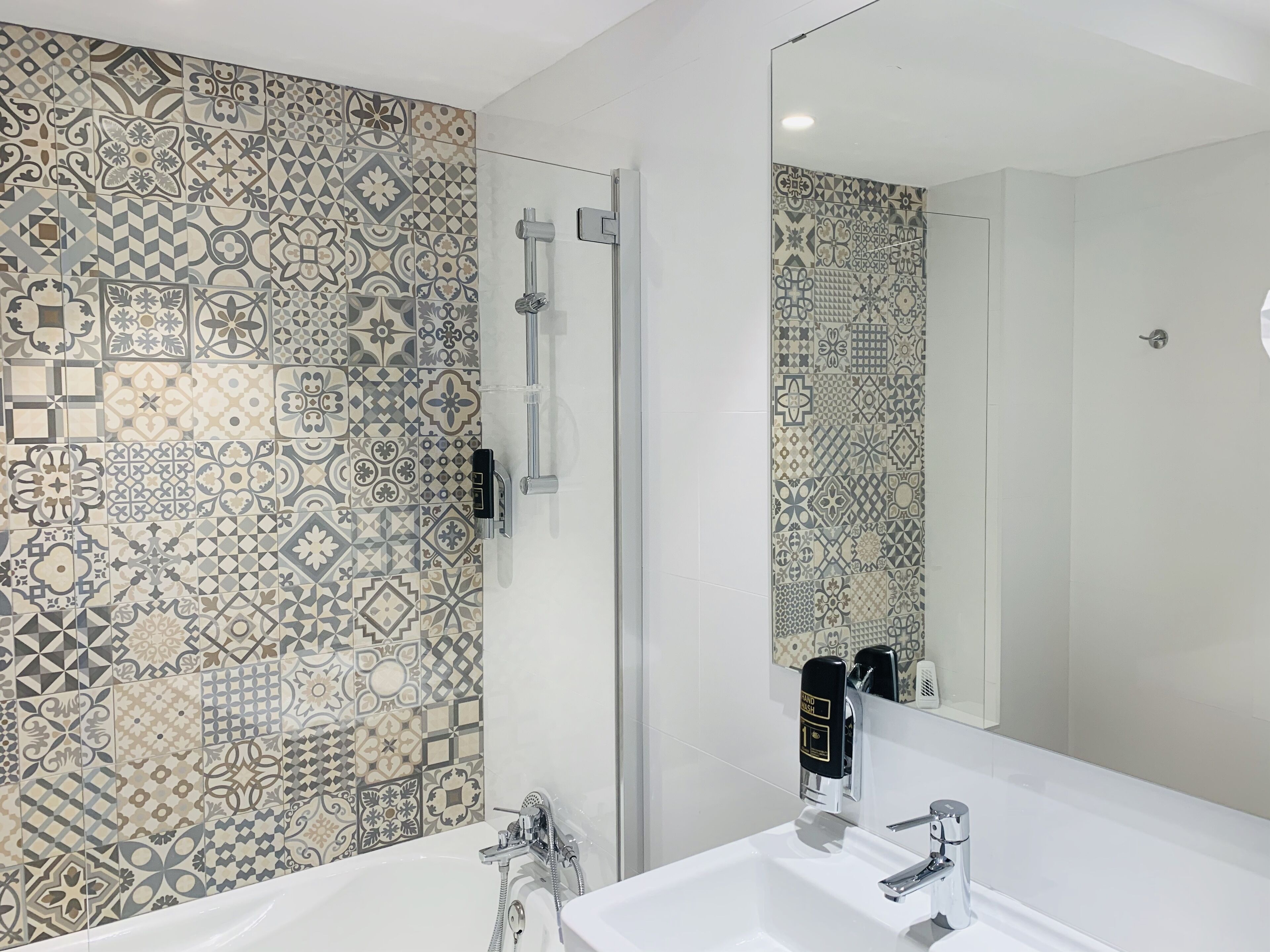 A bathroom with a patterned tile wall, a glass shower enclosure, a white sink, and a mirror.