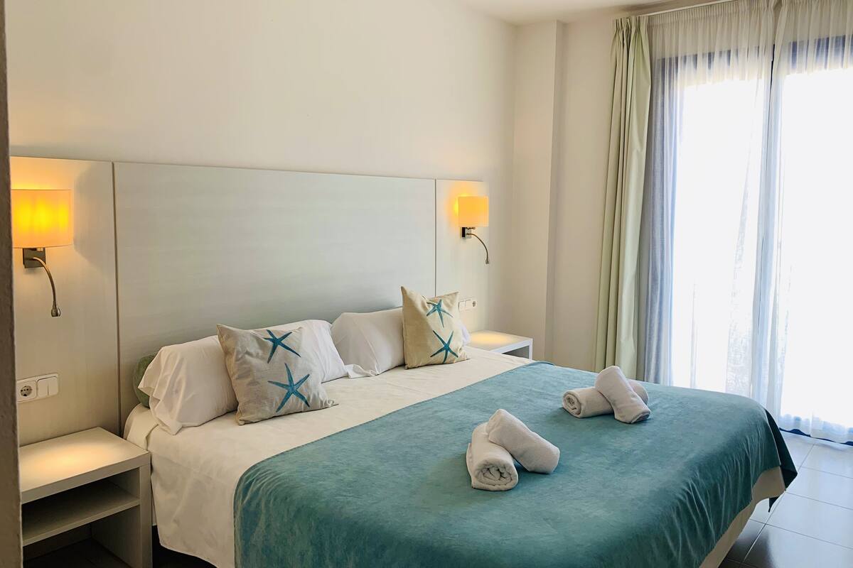 Una habitación de hotel con una cama, dos mesitas de noche, una lámpara y una ventana con cortinas.