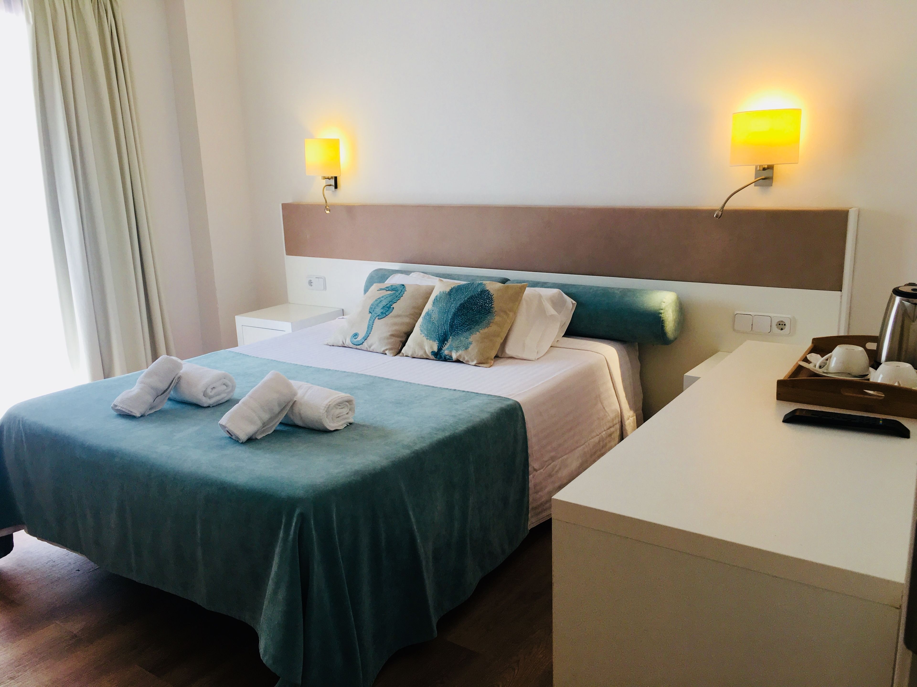 Habitación de hotel con cama, dos lámparas de noche, mesita de noche, escritorio y bandeja con tazas y tetera.