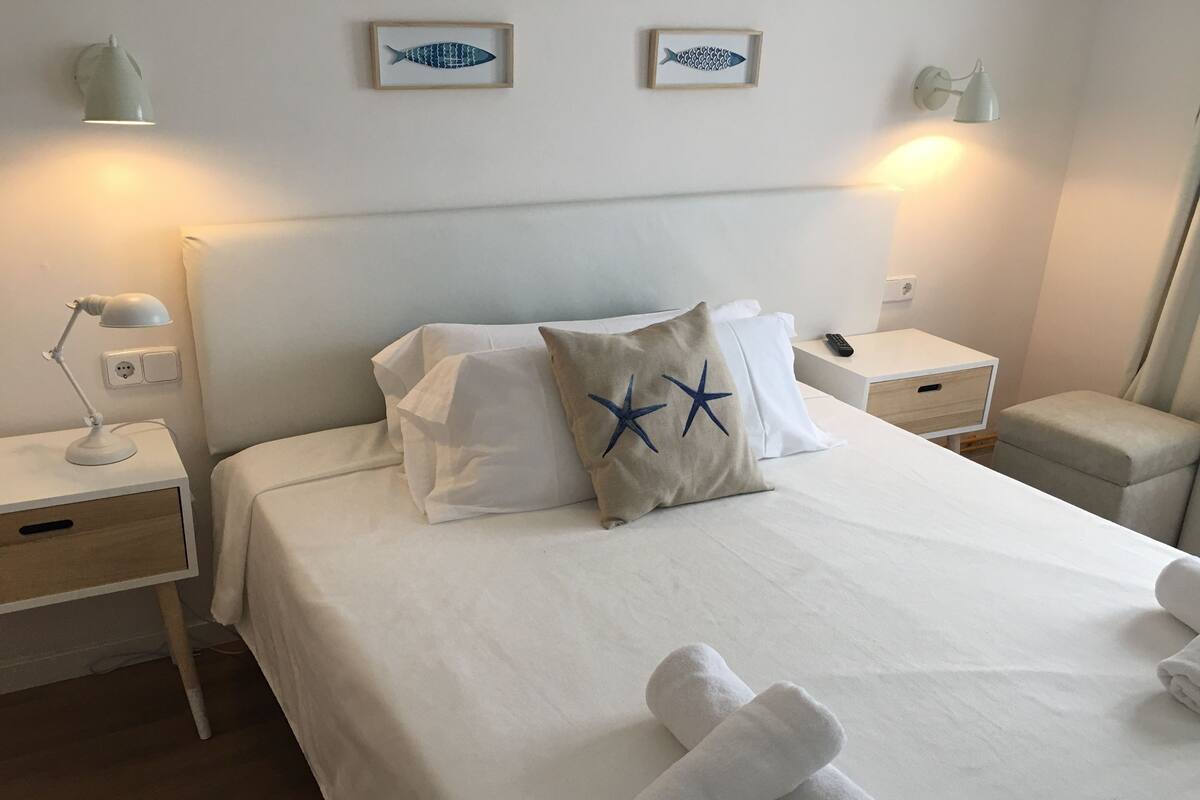 Una cama bien tendida con sábanas blancas, dos almohadas con forma de estrella de mar y toallas enrolladas sobre la cama. Hay dos mesitas de noche con lámparas y un mueble con un control remoto.