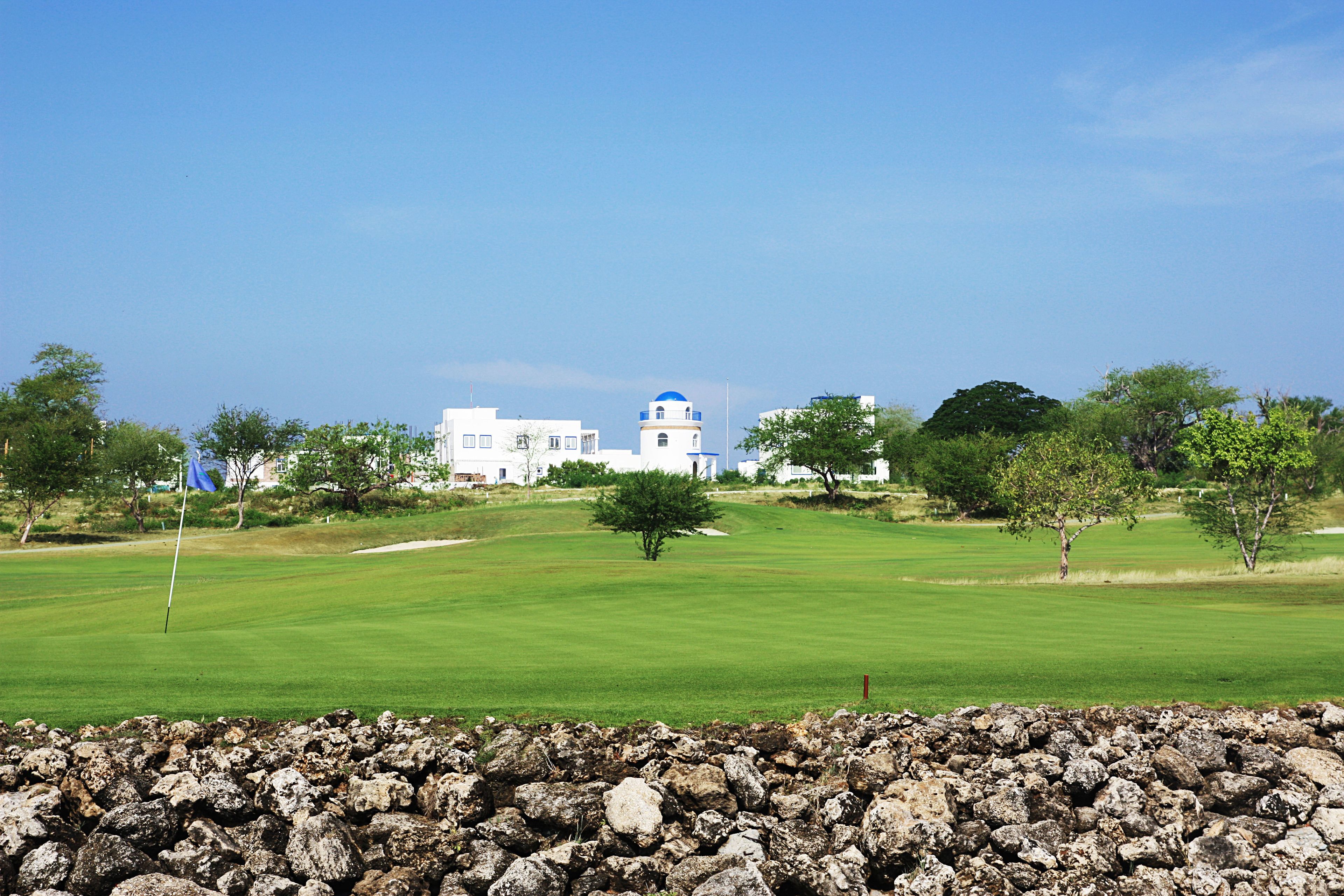 A golf course with a stone wall in the foreground, a clear blue sky, and a building with a blue dome in the background.
