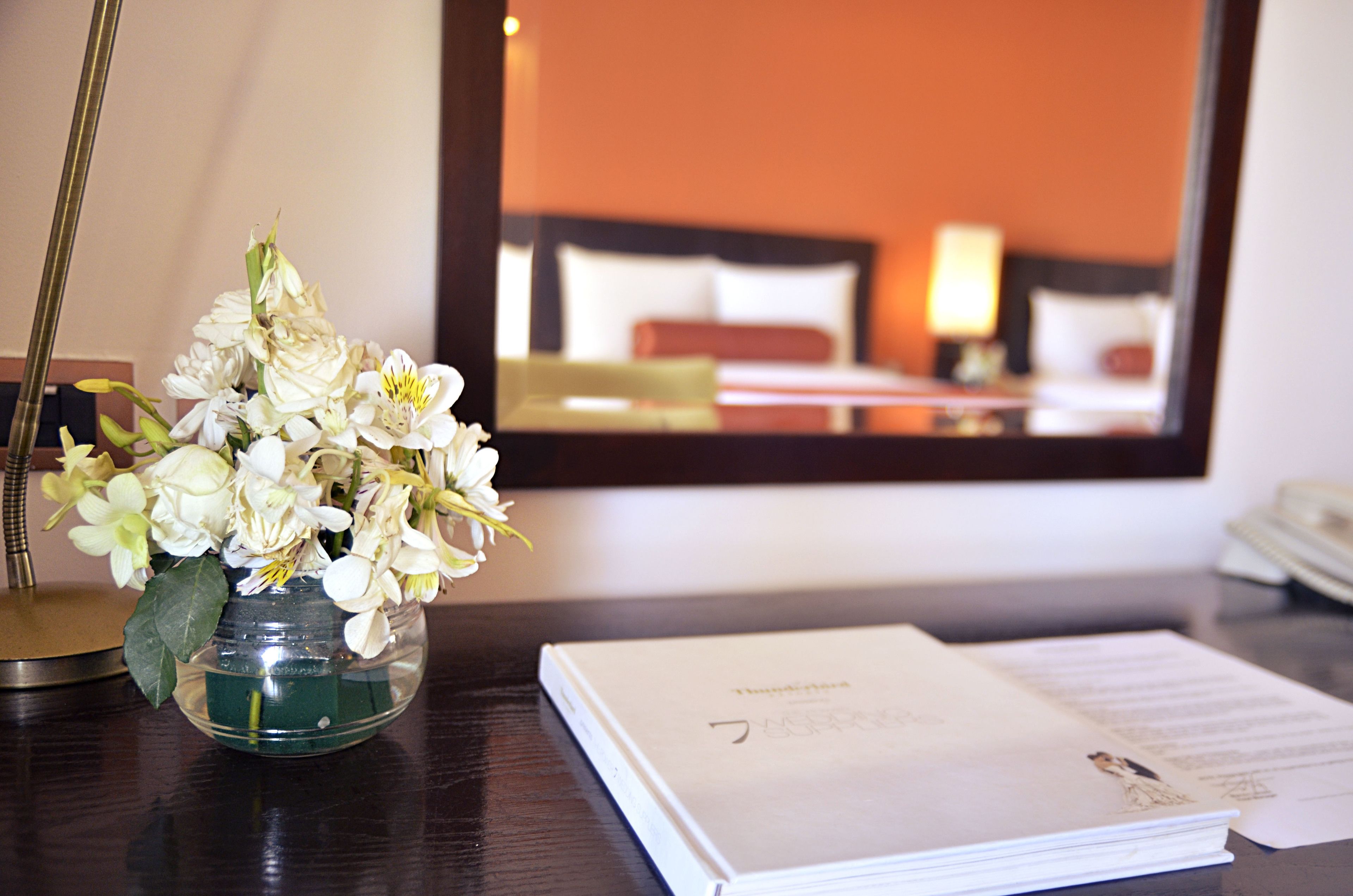 A hotel room with a bed, a bedside table with a book and papers, and a vase of flowers.