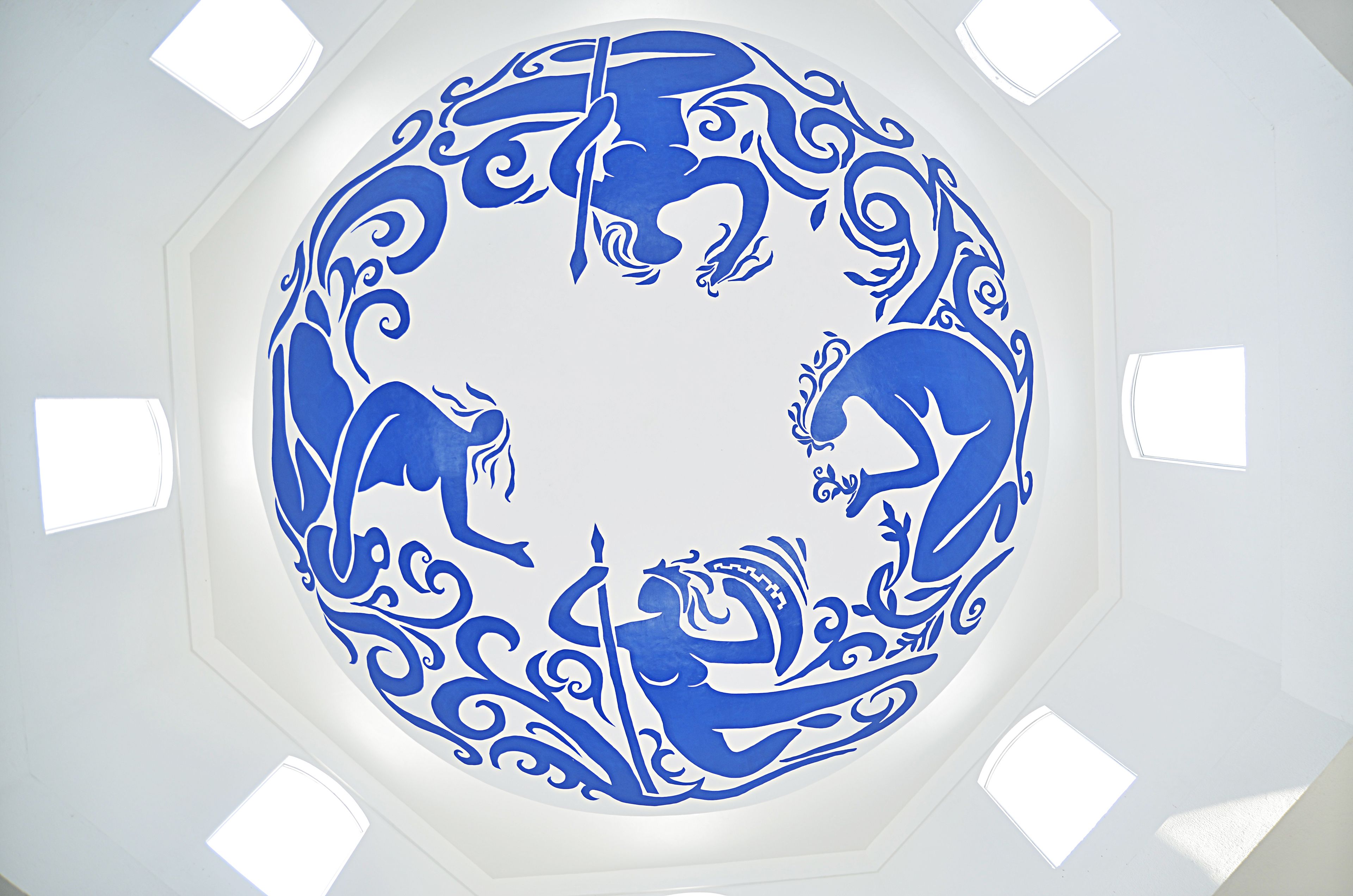 Een blauwe, ronde kunstinstallatie op een wit plafond, omringd door vier kleine ramen.
