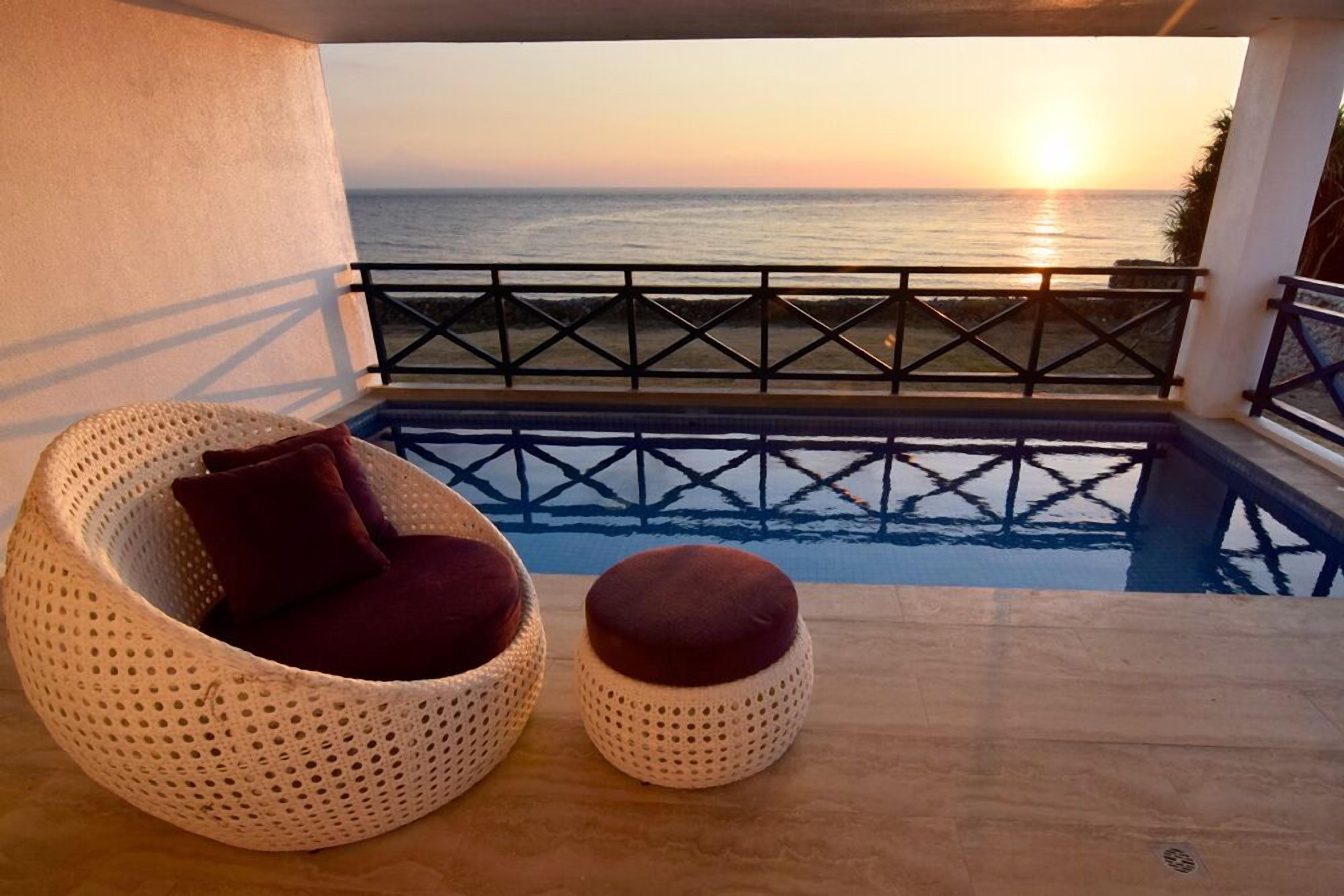 A balcony with a view of the sunset over the ocean, a wicker chair with a cushion, and a small ottoman.
