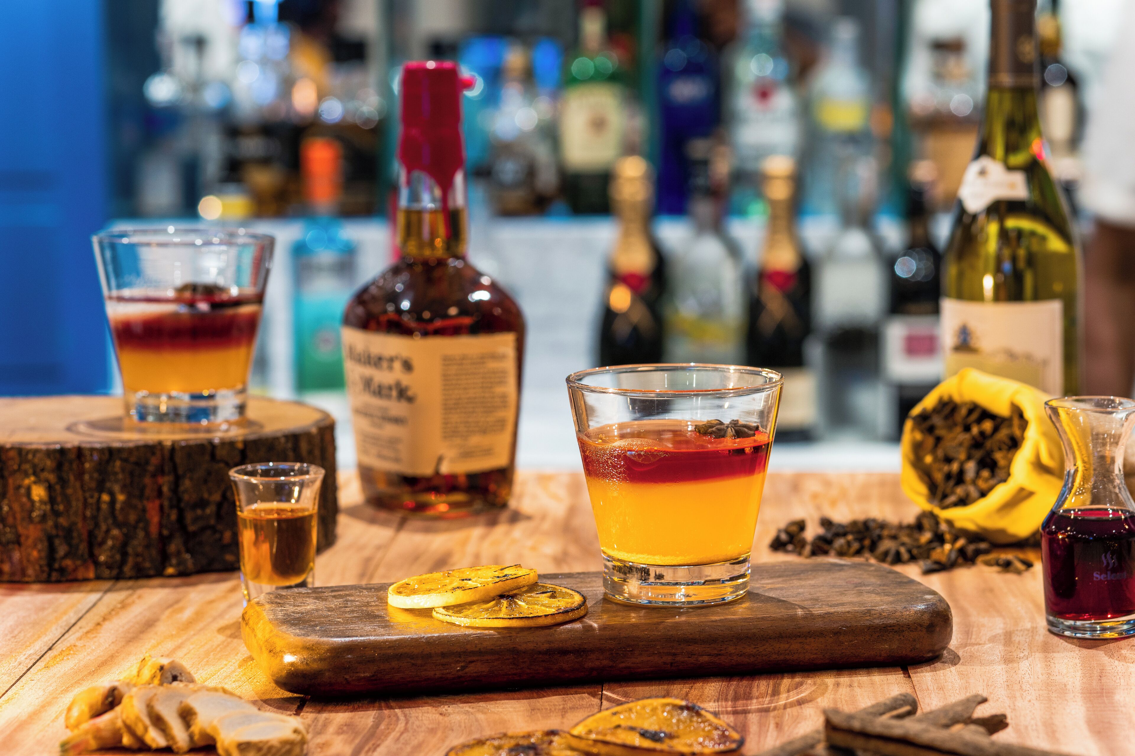 A bar setting with glasses of cocktails, a bottle of whiskey, and a wooden board with dried fruits.