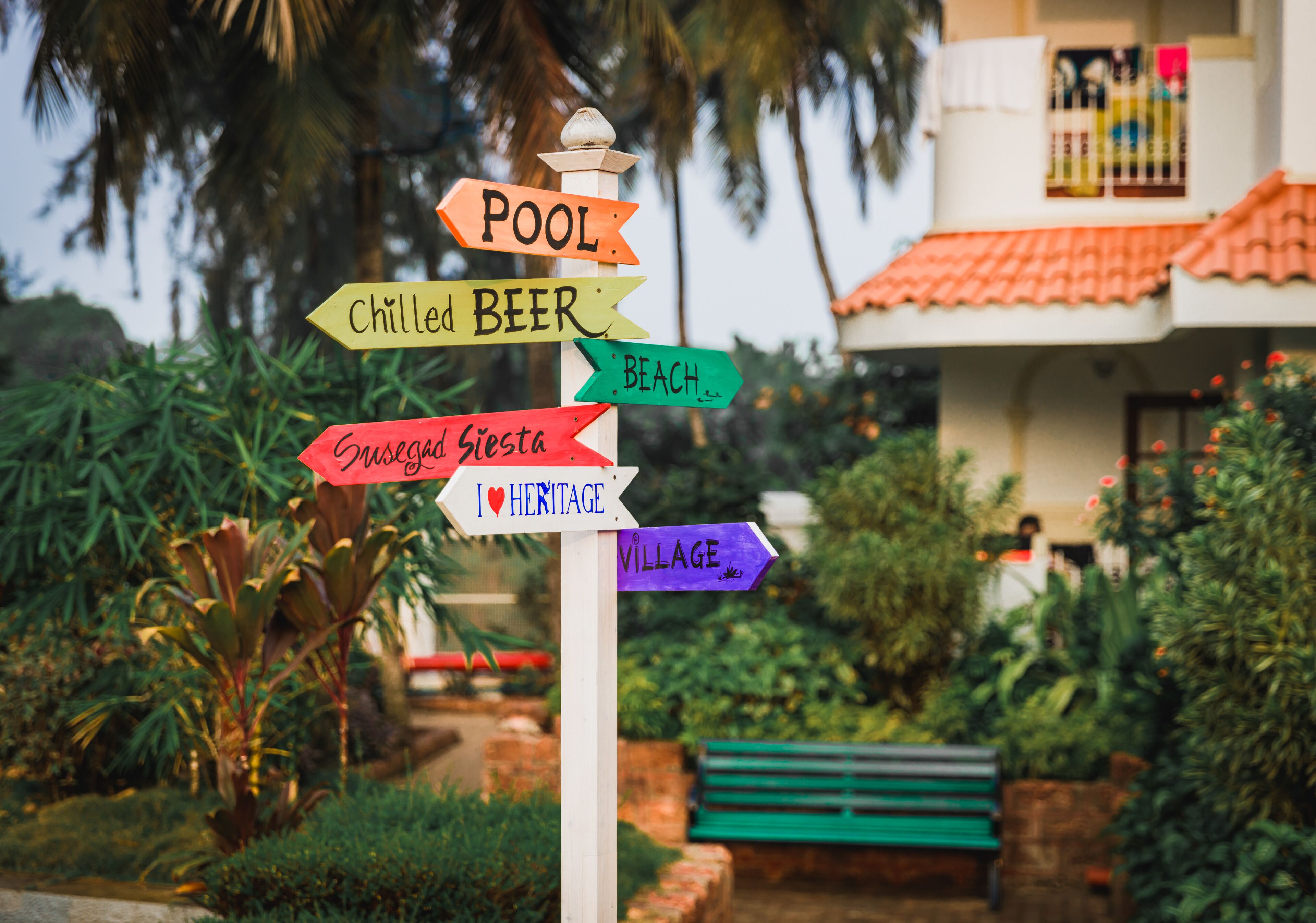 A signpost with multiple directional arrows pointing to various amenities such as a pool, chilled beer, beach, and village.