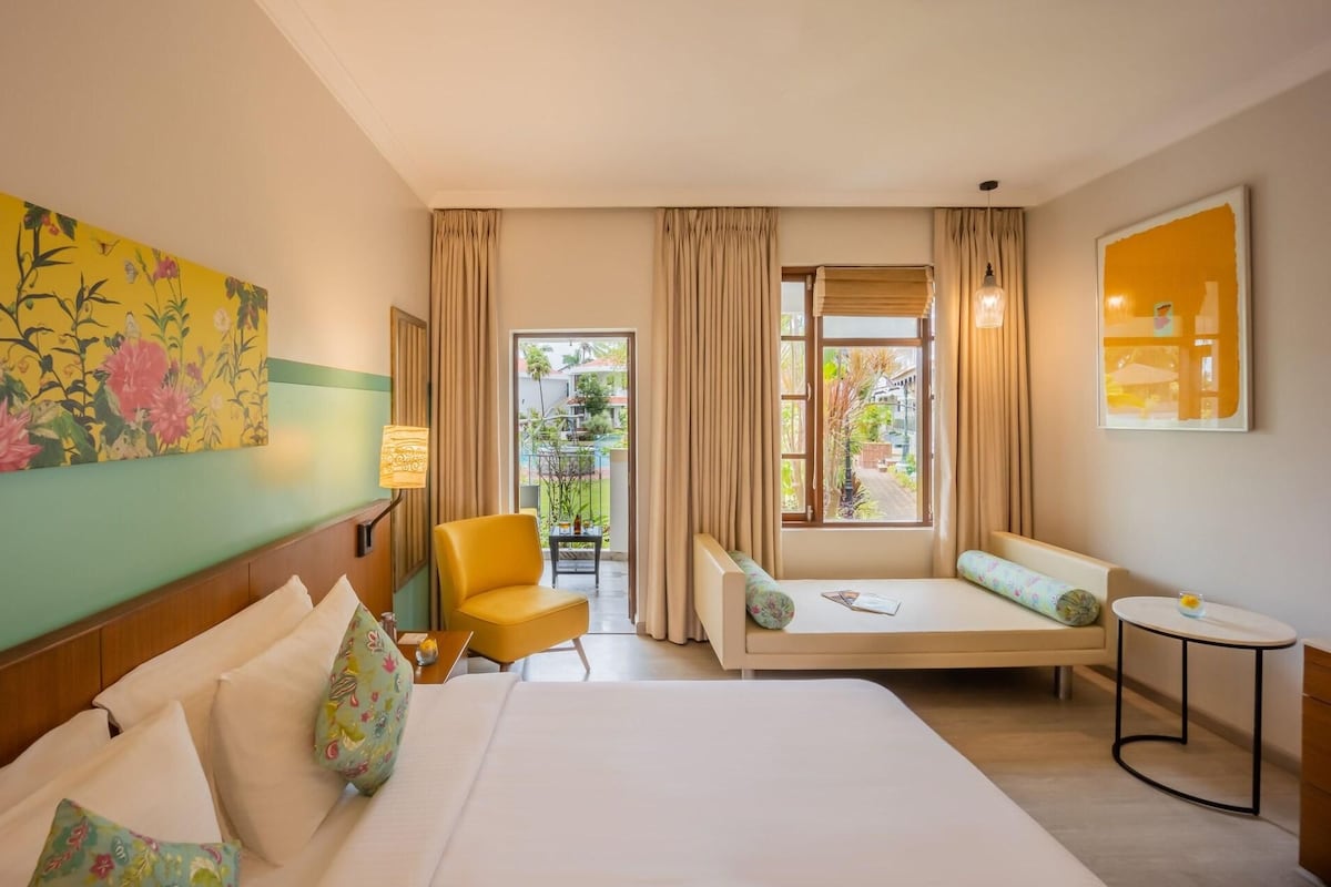 A hotel room with a bed, a sofa, a small table, a chair, and a balcony with curtains.