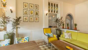 Een woonkamer met een groene bank, een gele fauteuil en een houten salontafel. Er staan potplanten, er hangen afbeeldingen in lijstjes aan de muur en de vloer heeft een patroon.