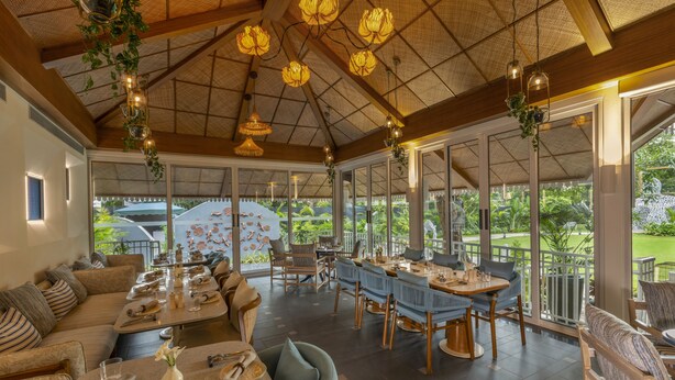 A spacious indoor dining area with high ceilings, large windows, and hanging floral decorations. - Heritage Village Resort & Spa Goa (Cansaulim)