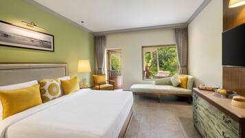 A hotel room with a large bed, a sofa, a TV, and a view of greenery outside.