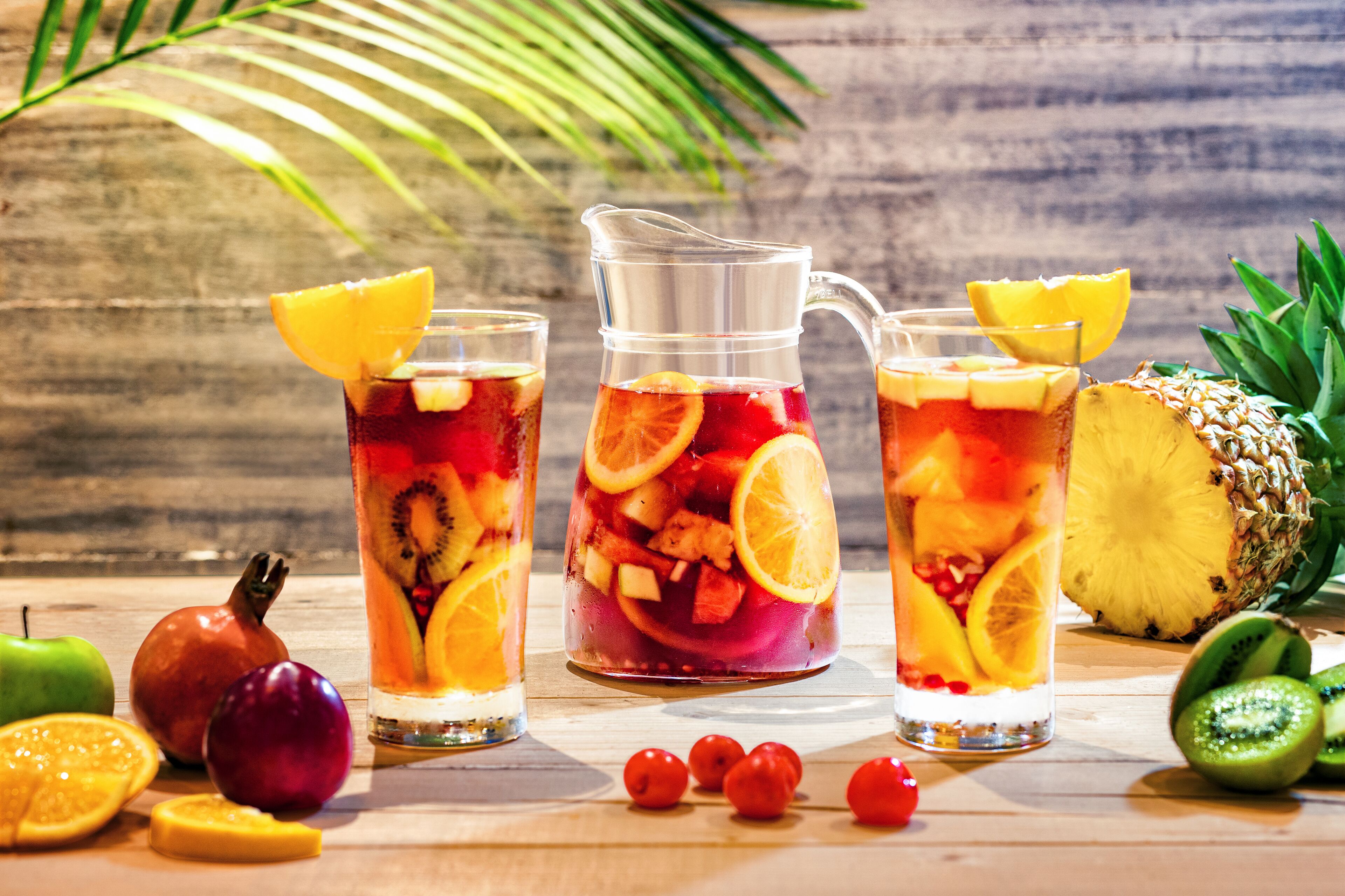 A wooden table with a pitcher and glasses of fruit-infused drinks, surrounded by fresh fruits and a pineapple.