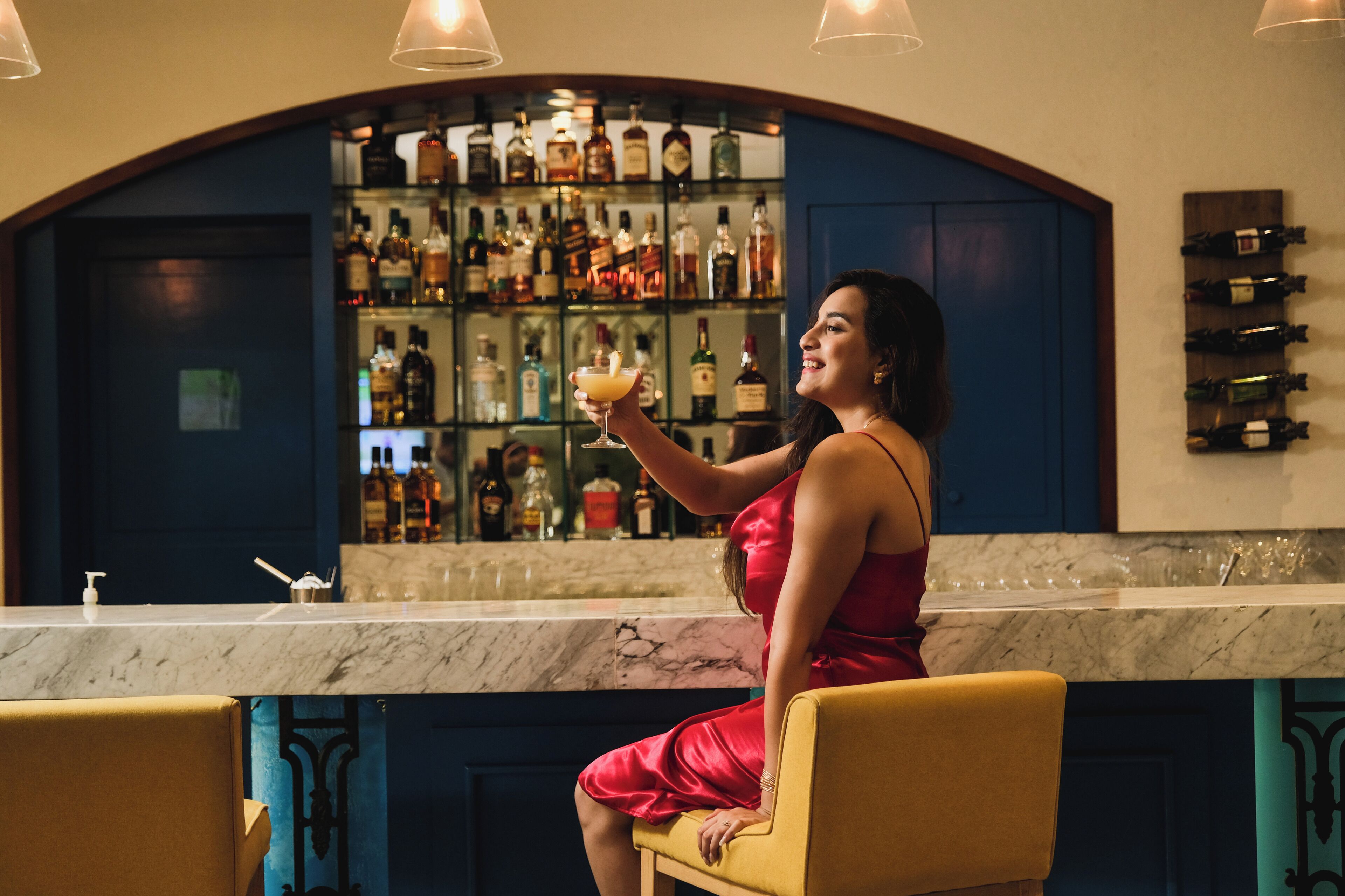 Mulher de vestido vermelho sentada num bar com uma bebida, rodeada por prateleiras de garrafas de licor.