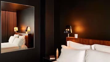Une chambre d’hôtel avec un grand miroir, un lit avec du linge de maison blanc, une table de chevet avec une lampe et une tête de lit en bois.